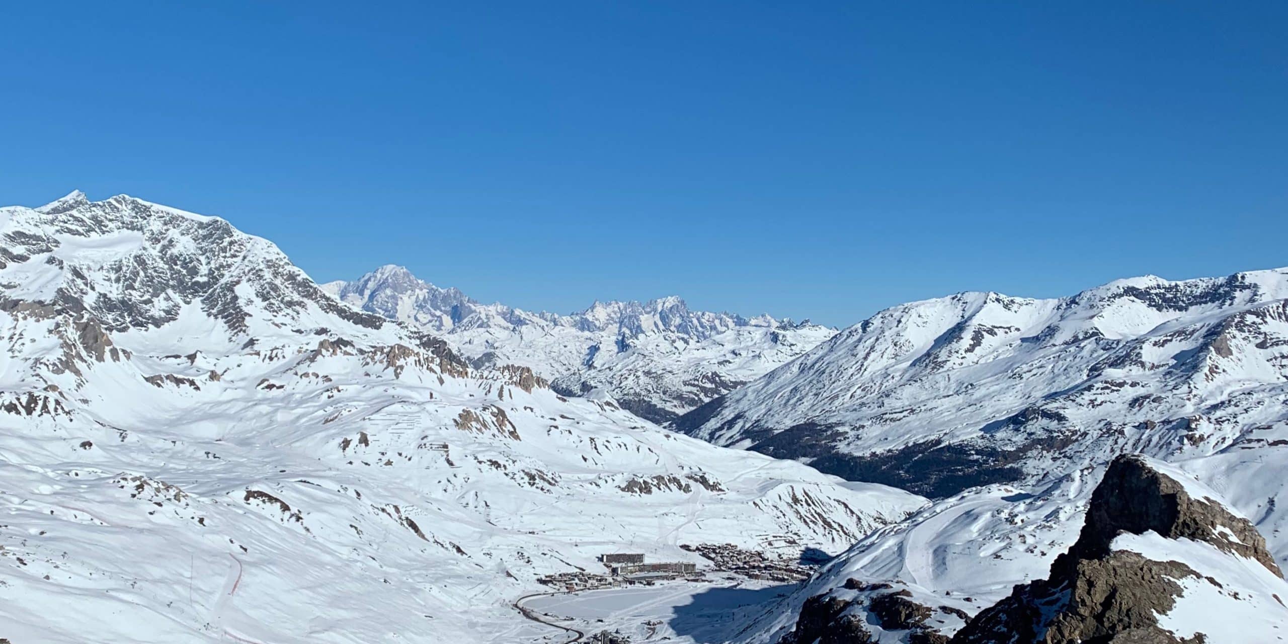 Paysage montagnard enneigé dominé par de hauts sommets et le Mont Blanc lointain. Un village de ski s'étend dans la vallée, sous un ciel d'un bleu éclatant.