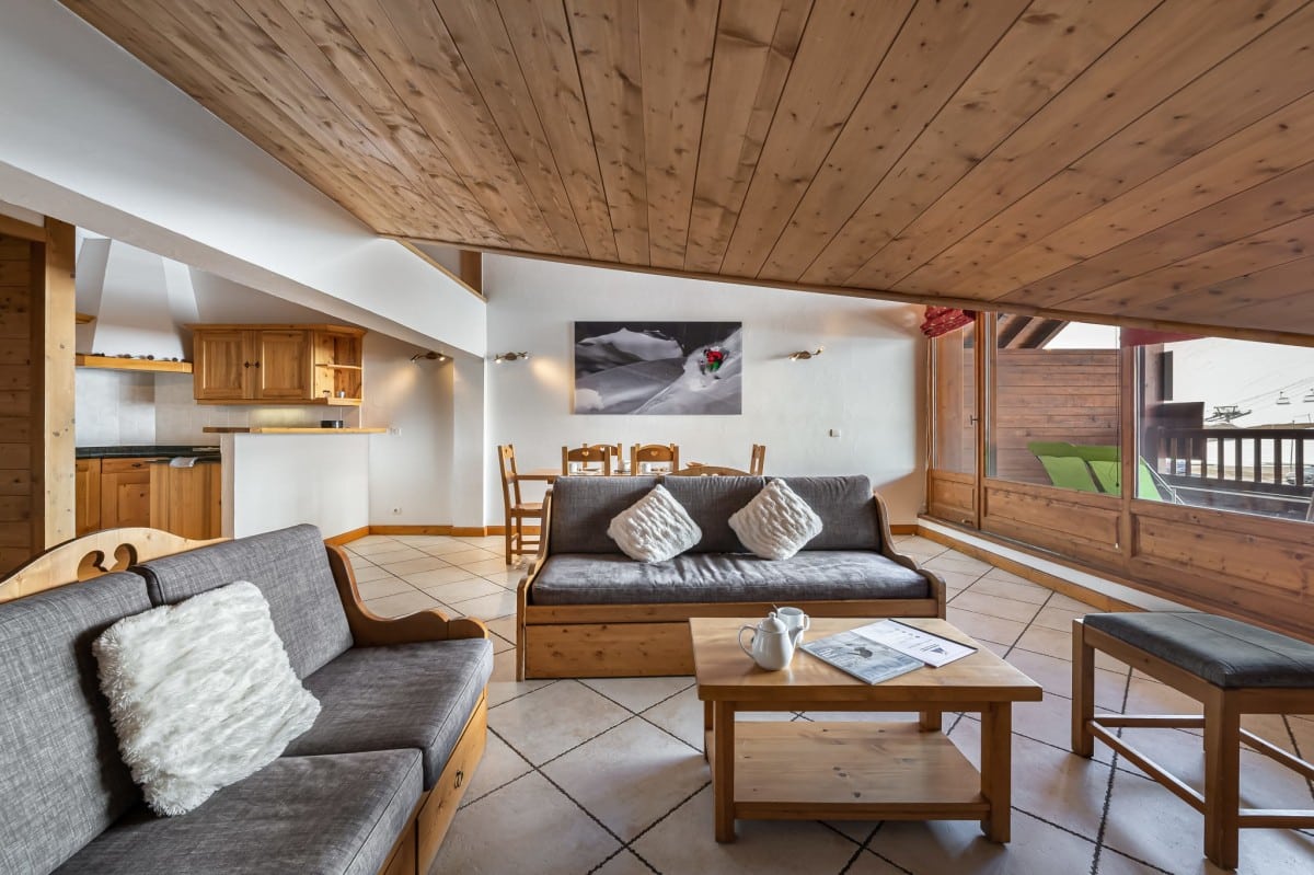 Grand salon-salle à manger-cuisine ouvert, aménagé avec des canapés et une table, donnant sur un balcon avec vue sur les télécabines, le tout sous un plafond en bois.
