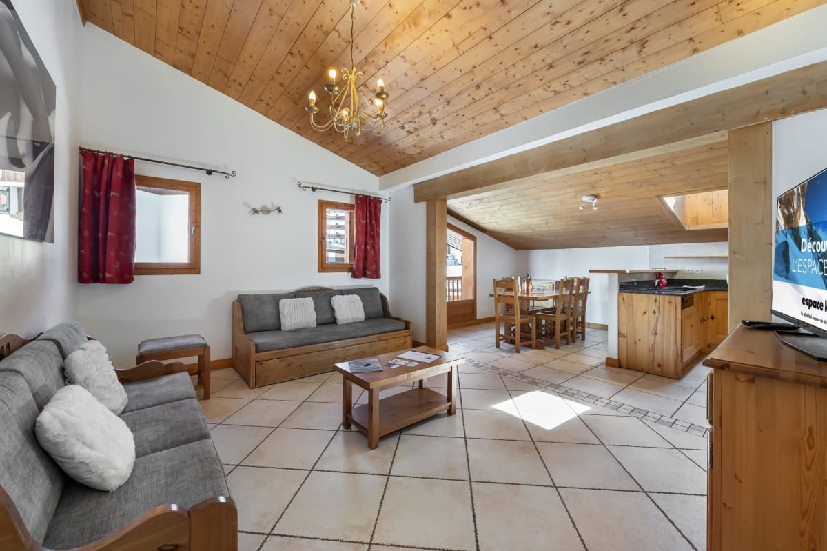 Un appartement de style chalet offre un espace de vie ouvert avec salon (canapés, table basse), salle à manger et cuisine. Plafonds en bois et sol carrelé. Sur la télévision : « Découv L'ESPACE espace ».