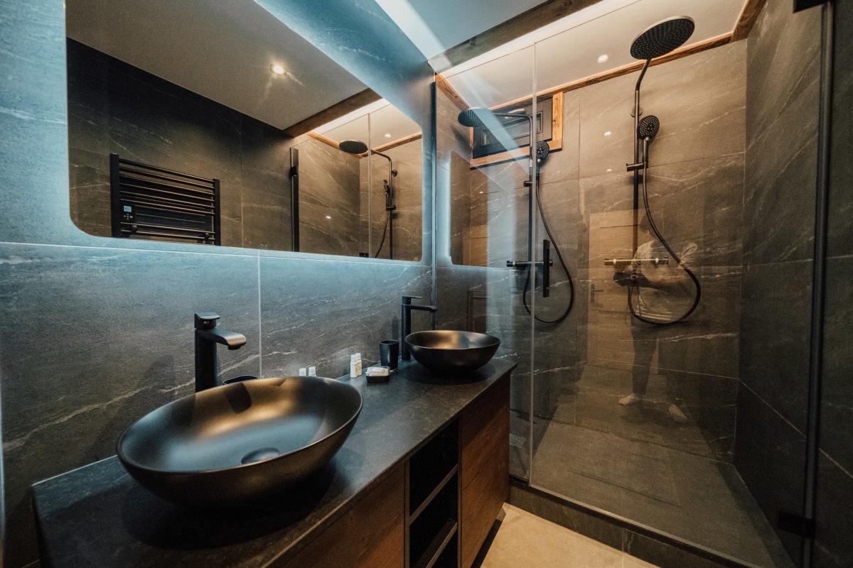Une salle de bain moderne présente deux vasques noires sur un meuble en bois foncé, devant un miroir rétroéclairé, avec une douche à l'italienne carrelée de dalles sombres.