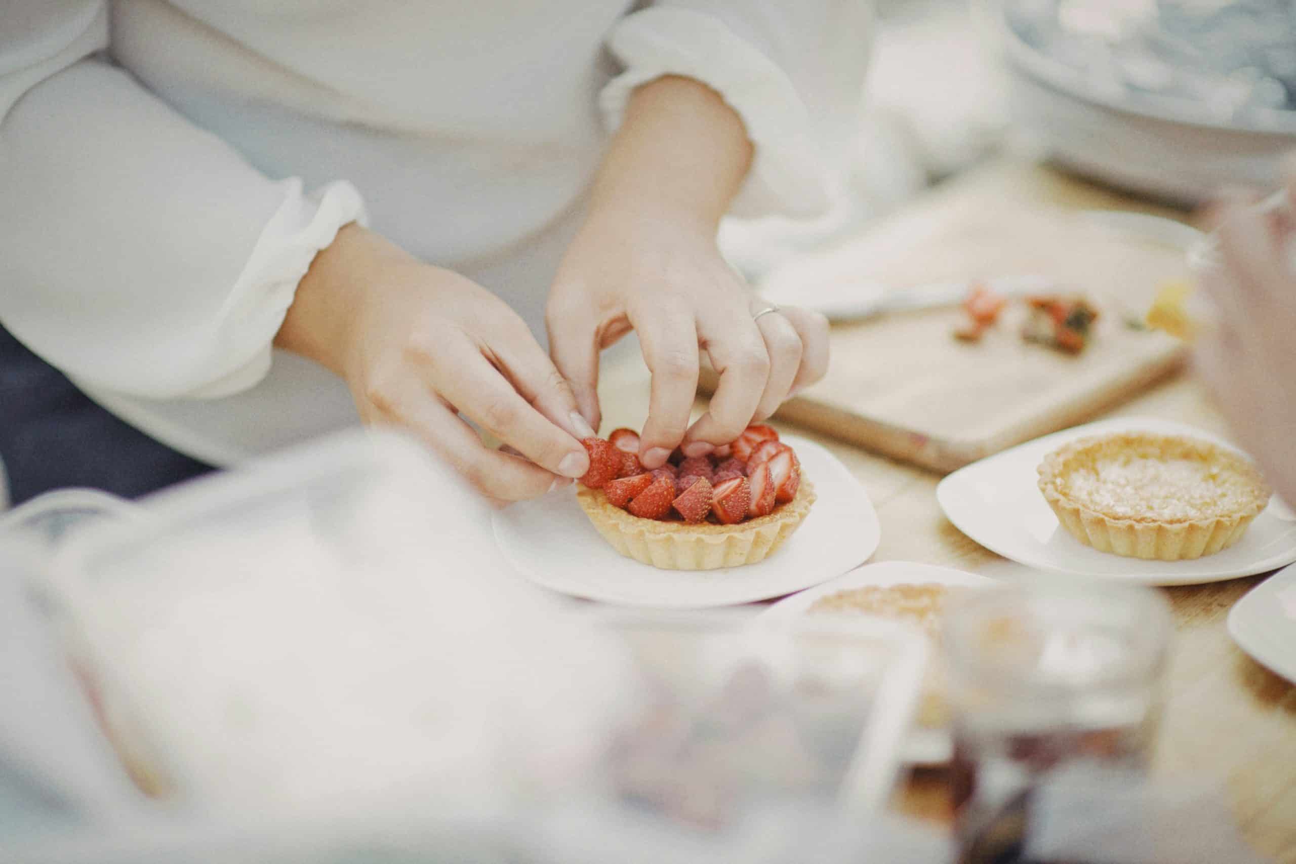 Des mains disposent des morceaux de fraises fraîches sur une tartelette garnie. La scène se déroule sur une table où d'autres pâtisseries et ustensiles de cuisine sont en arrière-plan.