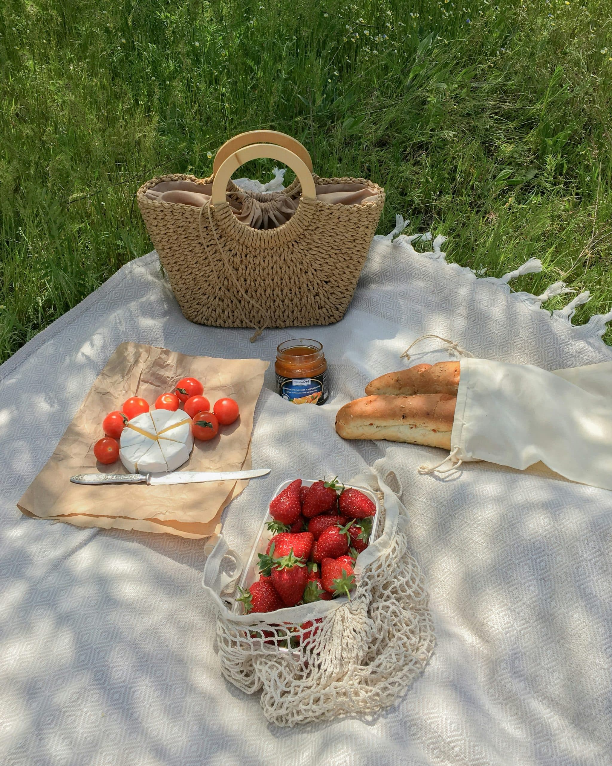 Un repas de pique-nique est étalé (fraises, camembert, tomates, baguettes, pot de confiture « HELCOM джем из абрикосов и мандаринов » et un sac en paille) sur une couverture claire dans l'herbe ensoleillée.
