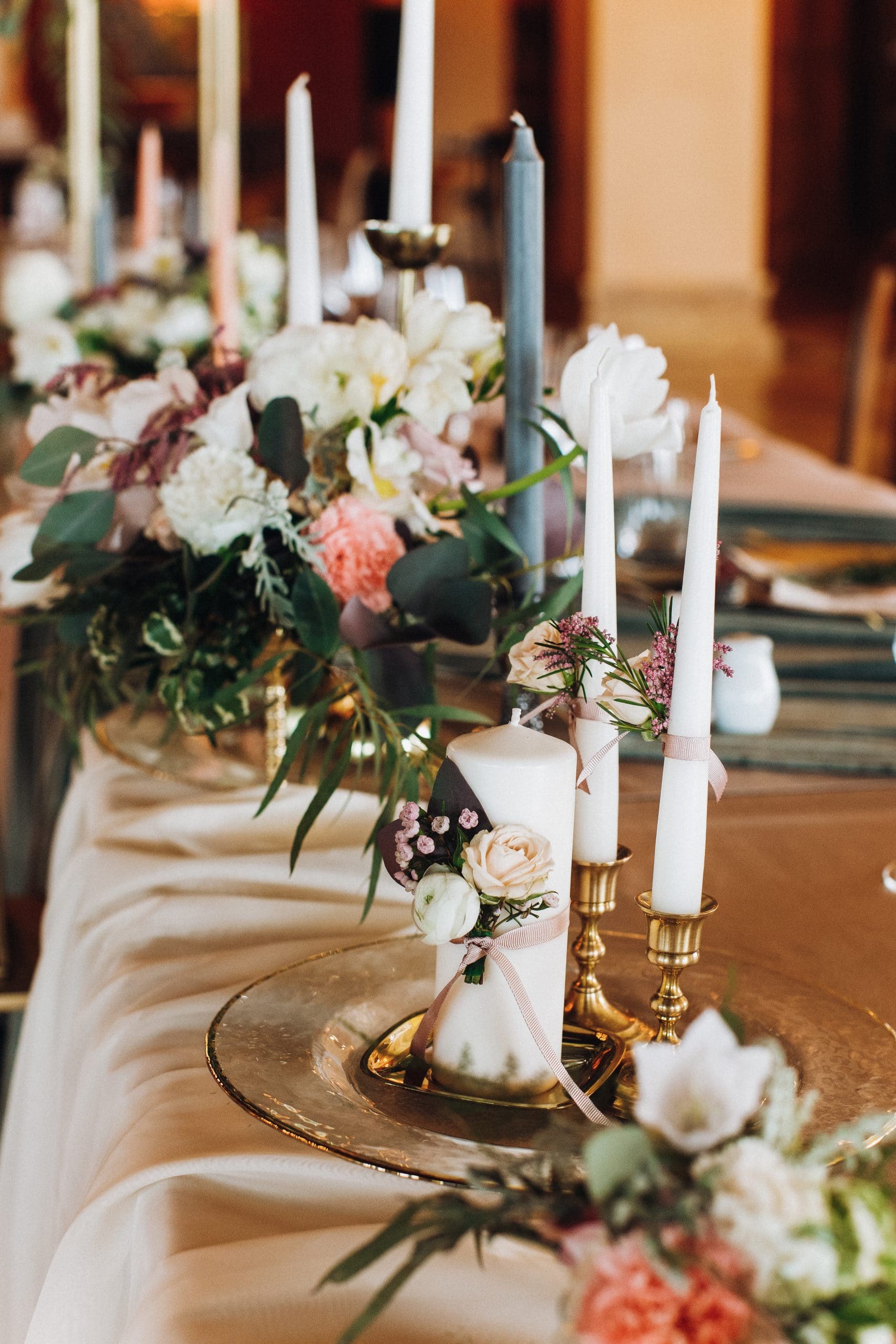 Bougies blanches et grises, décorées de rubans roses et de fleurs pastel, agrémentent une table de réception élégamment dressée avec des assiettes dorées et un centre floral.