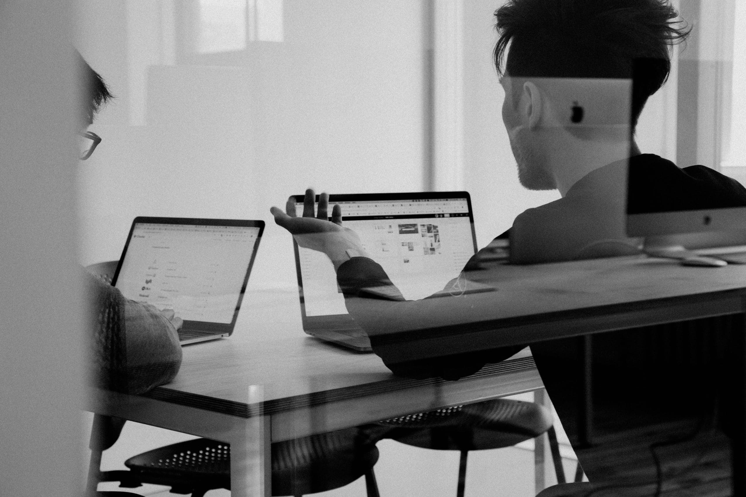 Deux personnes discutent et travaillent sur des ordinateurs portables. L'une, de dos, gesticule vers son écran. Scène de bureau en noir et blanc, vue à travers un verre réfléchissant.