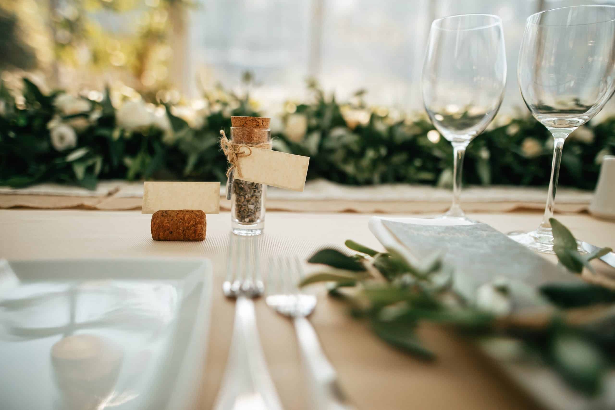 Une table de réception est dressée avec flacon (étiquette vierge), bouchon de liège, carte, vaisselle et verres. Décor floral et lumière douce créent une ambiance chaleureuse.