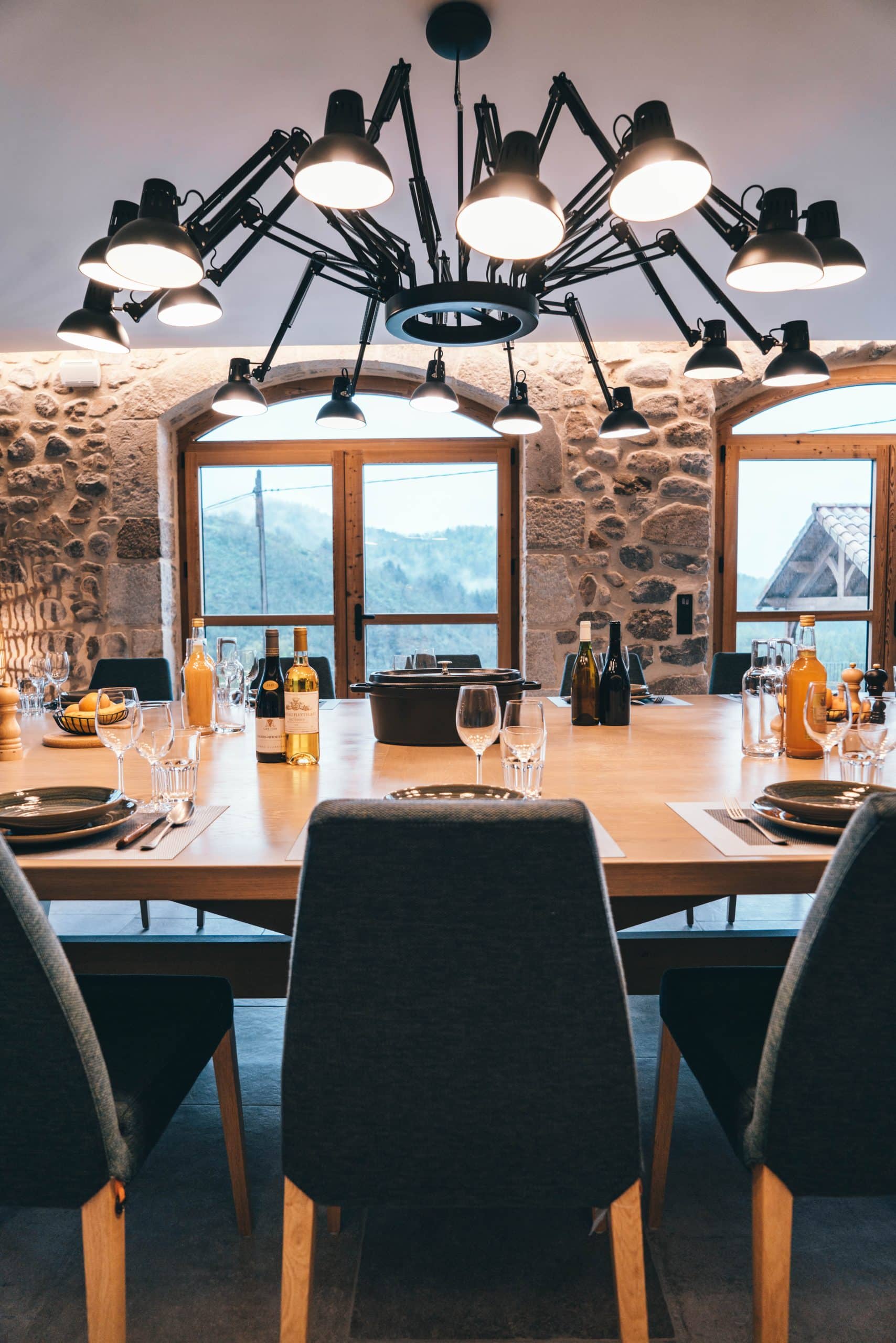 Une table en bois est dressée avec vaisselle, verres, bouteilles et une cocotte, entourée de chaises, sous un lustre industriel, dans une salle aux murs de pierre avec vue montagneuse. Texte sur les bouteilles : Bouteille rouge : "2019 GRAND CRU BURG FLEURTIGE APPELLATION D'ORIGINE CONTRÔLÉE MIS EN BOUTEILLE À LA PROPRIÉTÉ" Bouteille blanche : "BOURGOGNE FLEURTIGE APPELLATION D'ORIGINE CONTRÔLÉE MIS EN BOUTEILLE À LA PROPRIÉTÉ"