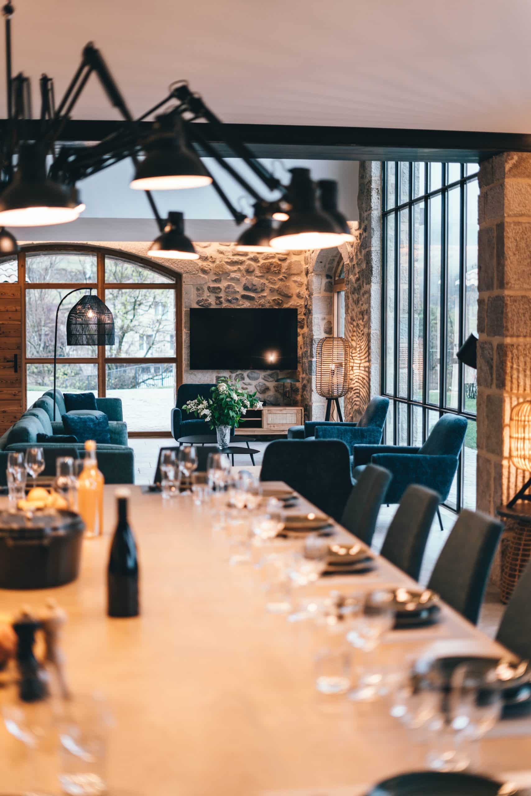 Une table de repas moderne, dressée avec verres et bouteilles, s'étend vers un grand salon avec des canapés bleus, des murs en pierre et de hautes baies vitrées. Des suspensions industrielles éclairent la scène.