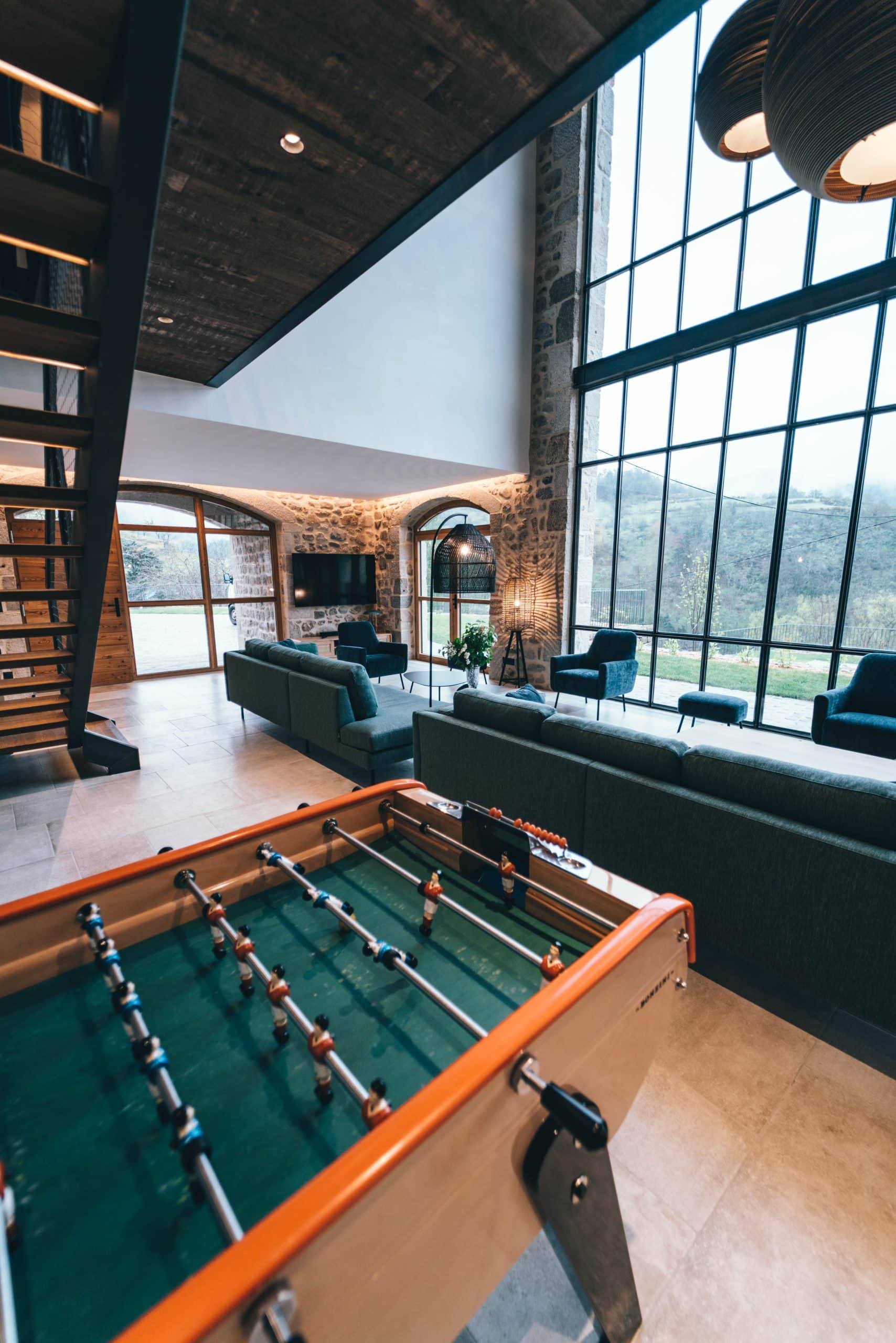 Un baby-foot BONZINI au premier plan d'un vaste salon moderne-rustique avec canapés, murs en pierre et grandes baies vitrées donnant sur une vallée.