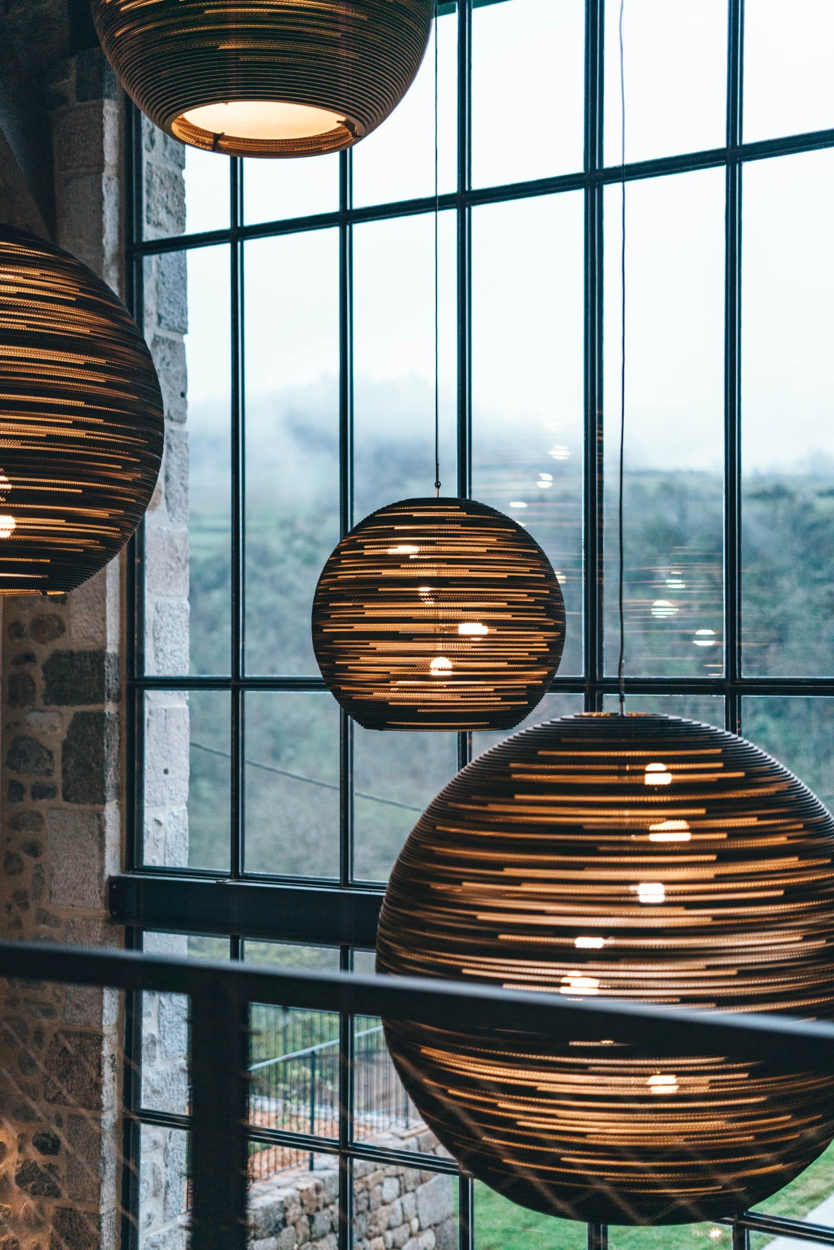 Trois luminaires sphériques en bandes de carton diffusent une lumière chaude devant une grande fenêtre, donnant sur un paysage verdoyant et brumeux, avec un mur de pierres apparentes sur la gauche.
