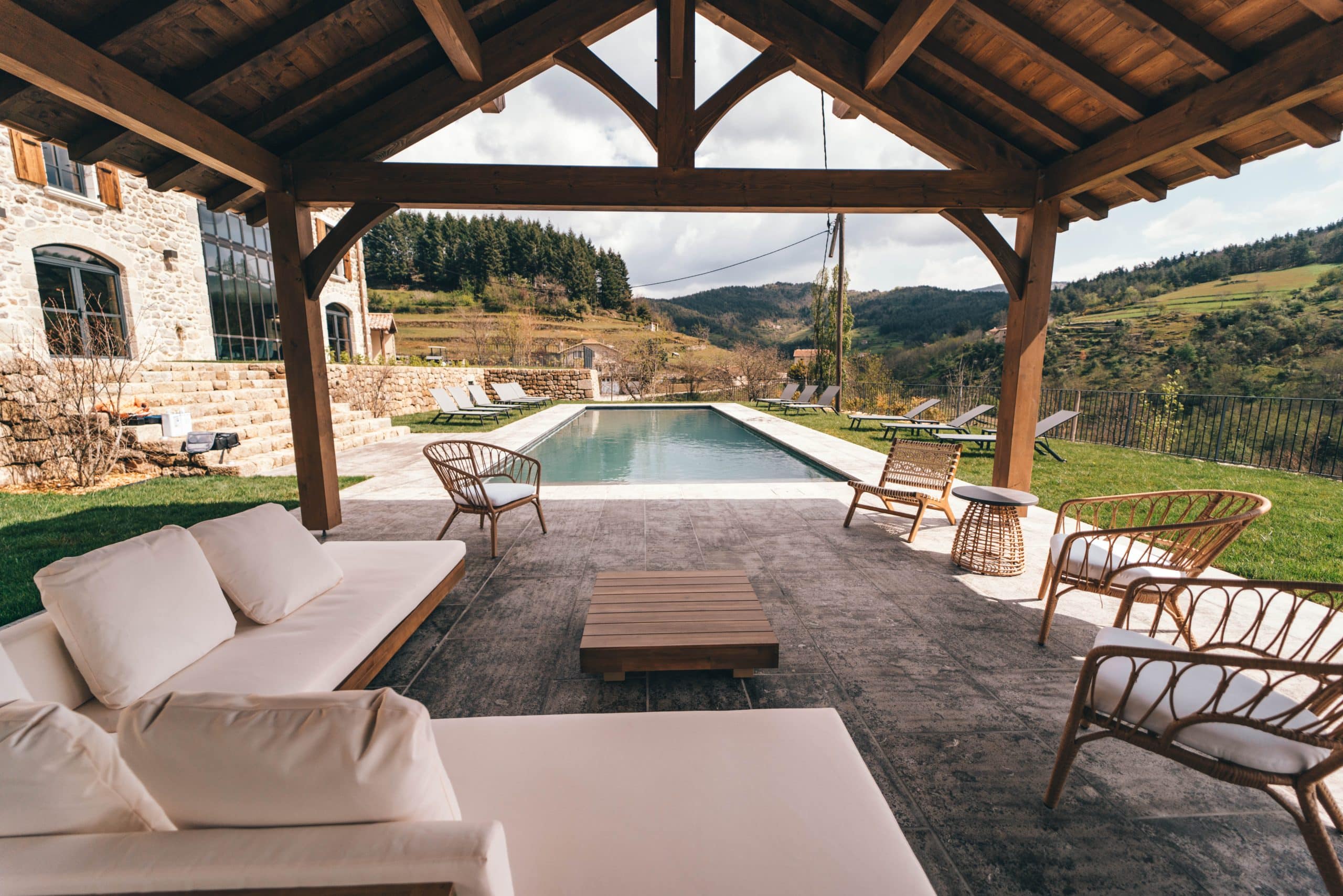Un espace détente moderne avec canapés et chaises sous une pergola en bois, jouxte une piscine et un bâtiment en pierre, offrant une vue sur une vallée verdoyante et des collines boisées.