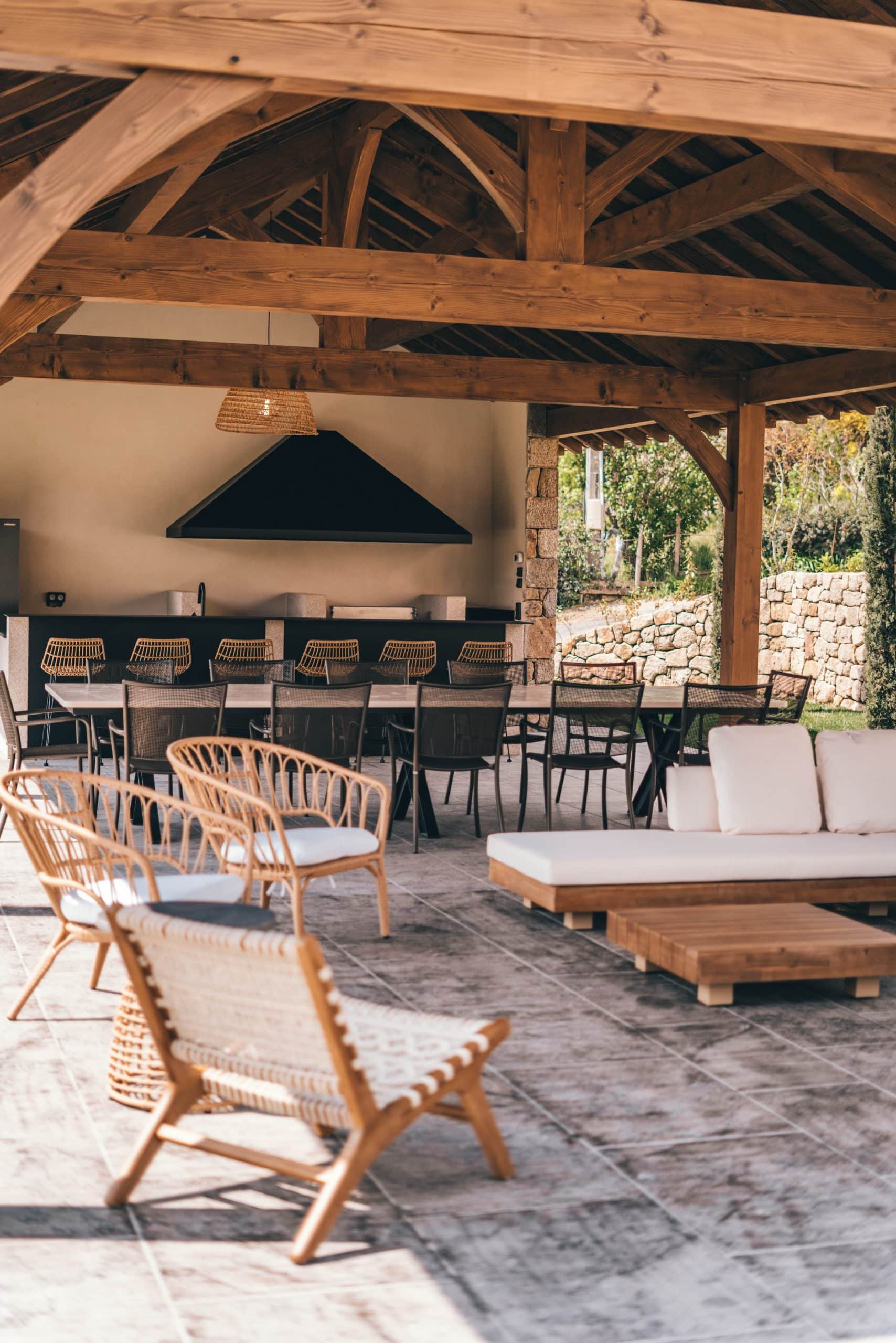 Un grand espace extérieur couvert par une pergola en bois abrite un coin repas et une cuisine d'été avec un salon attenant, devant un mur de pierres.