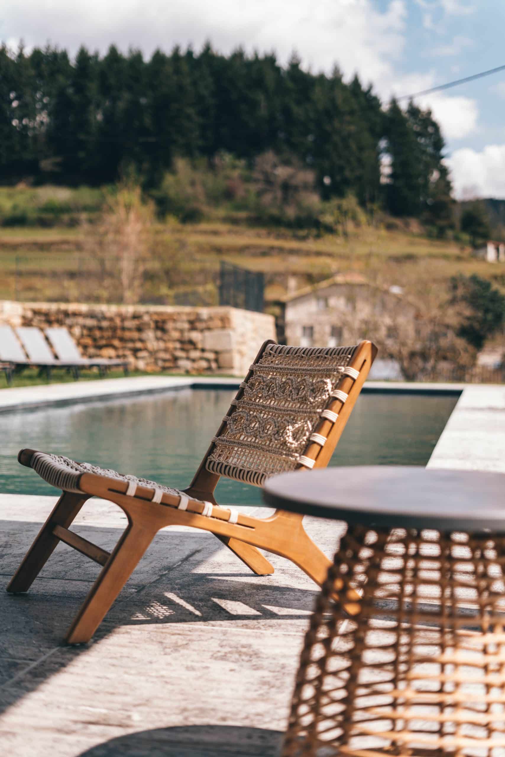Chaise longue en bois et macramé sur une terrasse carrelée, bordant une piscine. En arrière-plan, une colline boisée, une maison et un mur de pierres sous un ciel nuageux.