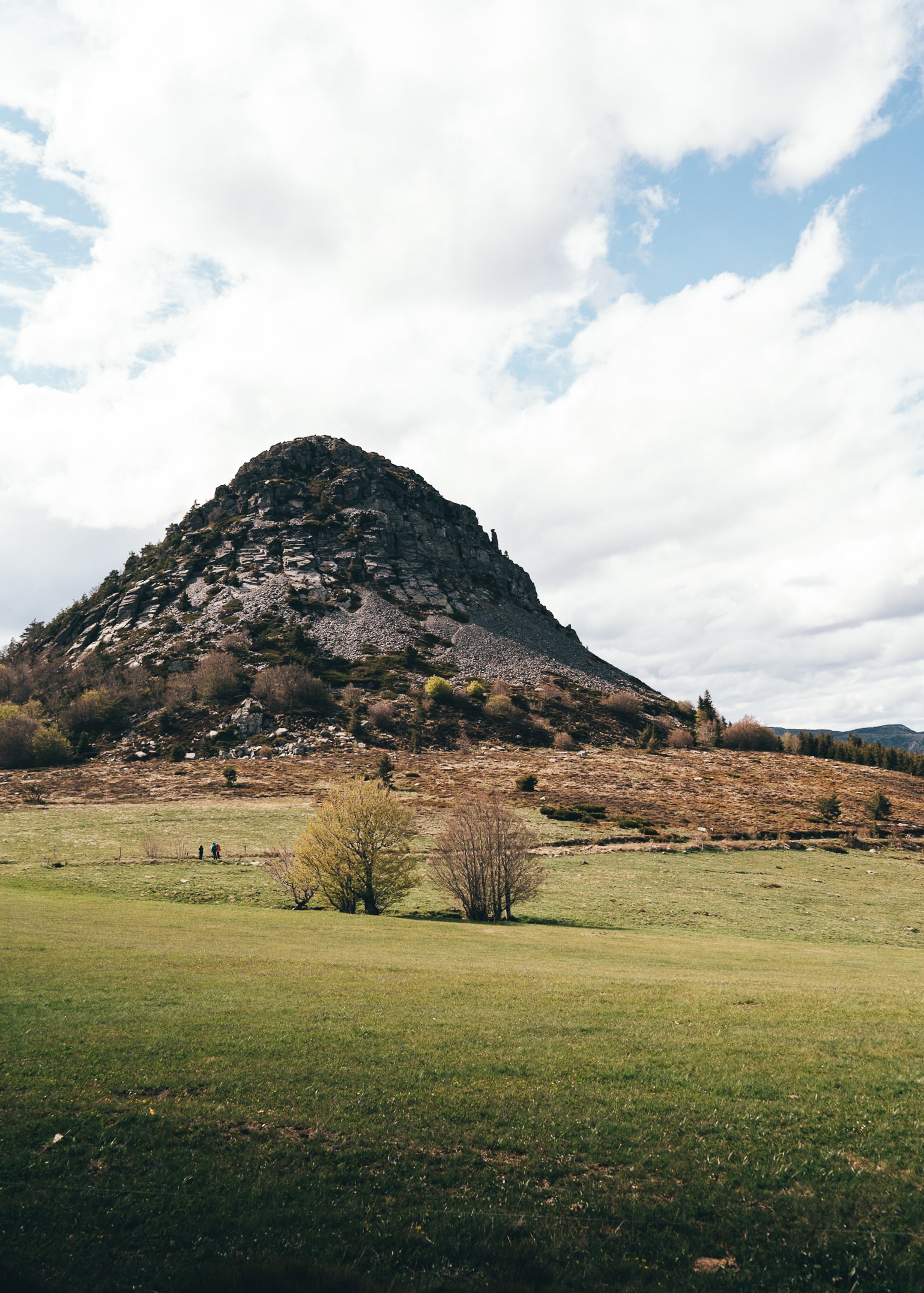 Une imposante montagne rocheuse domine un vaste pré verdoyant où deux randonneurs marchent. Le ciel est partiellement nuageux, avec quelques arbres épars.