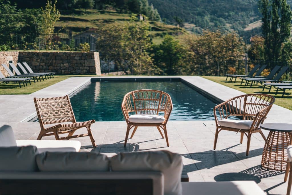 Trois chaises de salon en rotin et tressées, avec coussins, bordent une piscine rectangulaire sur une terrasse carrelée. Des transats sont sur la pelouse, devant un paysage de collines boisées sous le soleil.