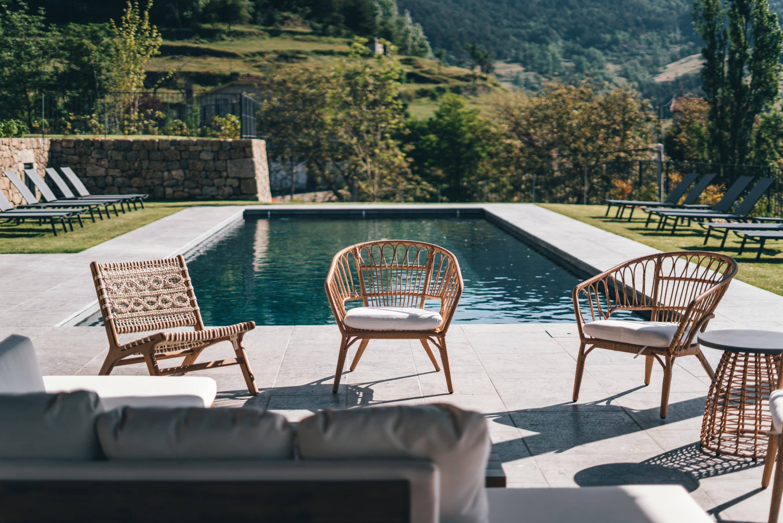 Trois chaises de salon en rotin et tressées, avec coussins, bordent une piscine rectangulaire sur une terrasse carrelée. Des transats sont sur la pelouse, devant un paysage de collines boisées sous le soleil.