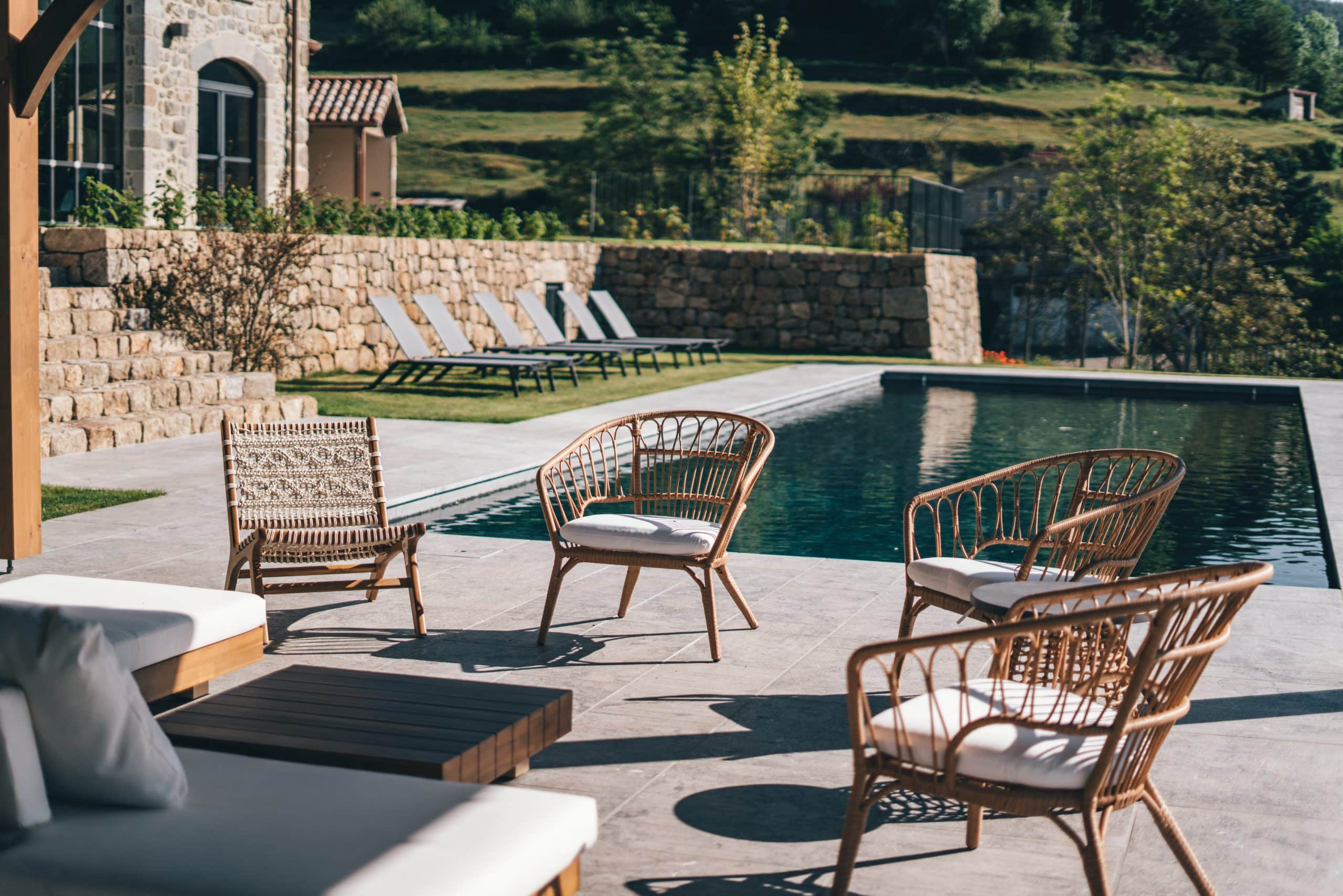 Fauteuils en rotin et canapé blanc invitent à la détente sur une terrasse moderne. Une piscine rectangulaire miroite, bordée par une maison en pierre et des collines verdoyantes.