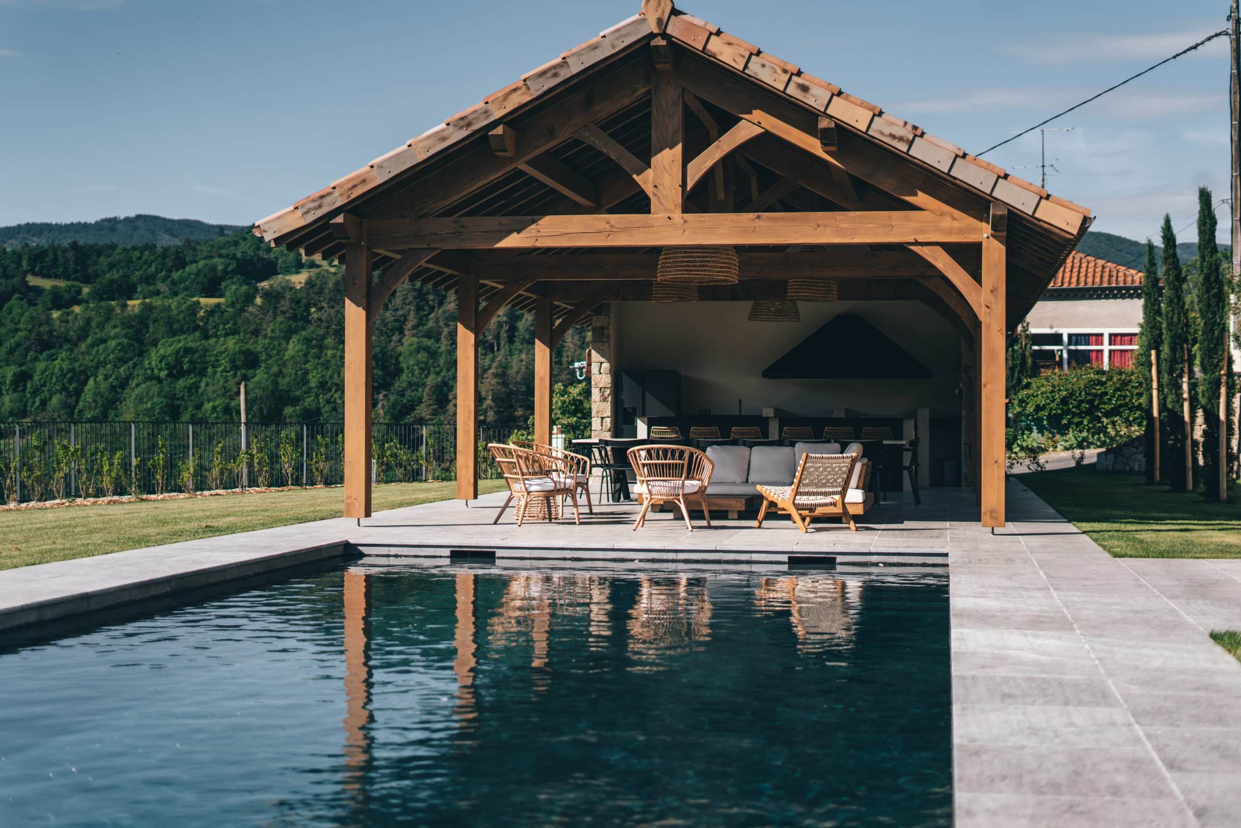 Grande pergola en bois abritant un salon de jardin et une cuisine d'été, située au bord d'une piscine sombre, face à des collines verdoyantes sous un ciel bleu.