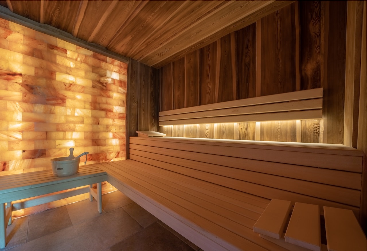 Un sauna moderne offre un espace de détente avec des bancs en bois clair, un mur lumineux de briques de sel roses et un seau "HARVIA".