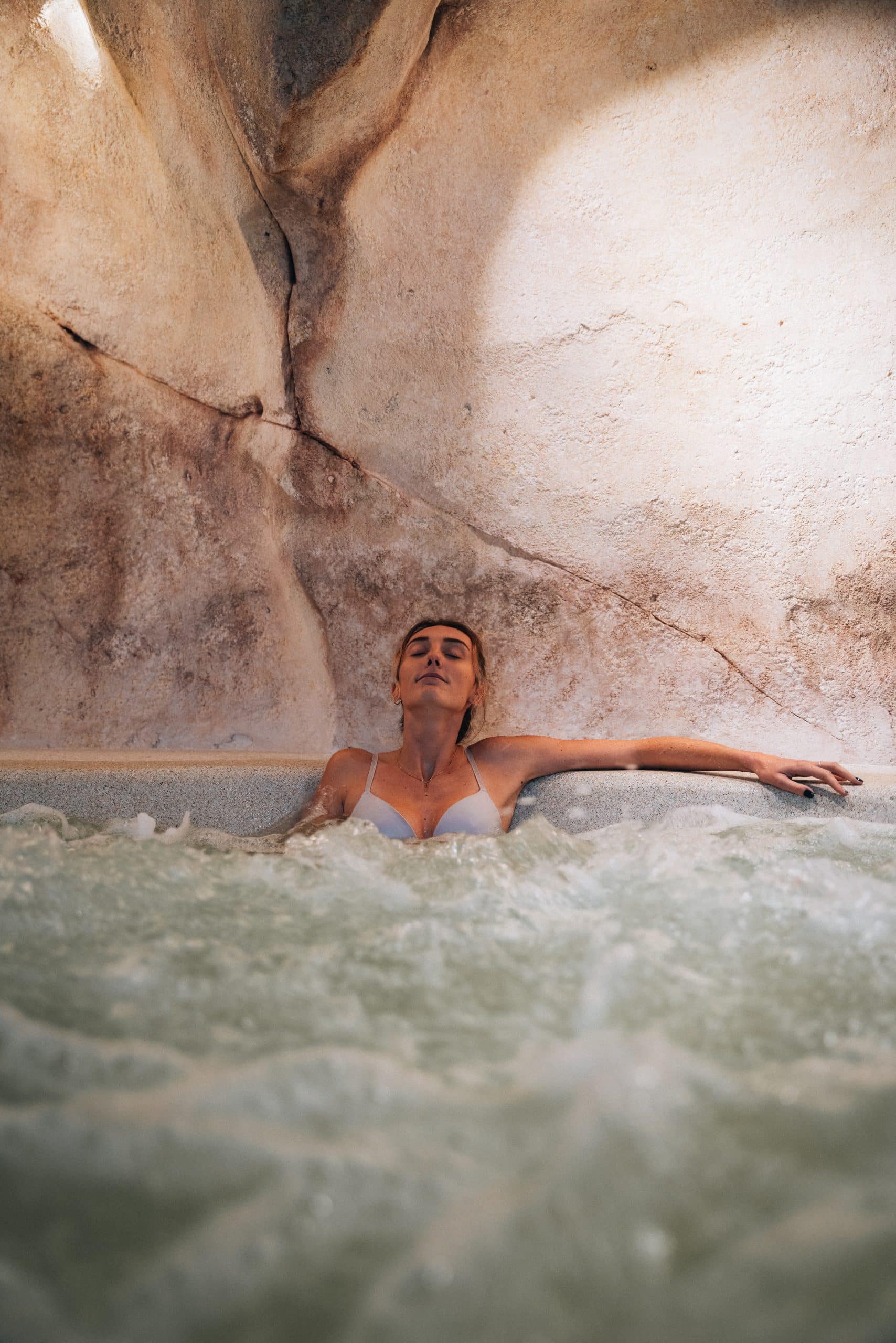Une femme aux yeux clos se détend dans un bain à remous bouillonnant, adossée à des parois en pierre claires. Son bras droit est posé sur le rebord du spa.