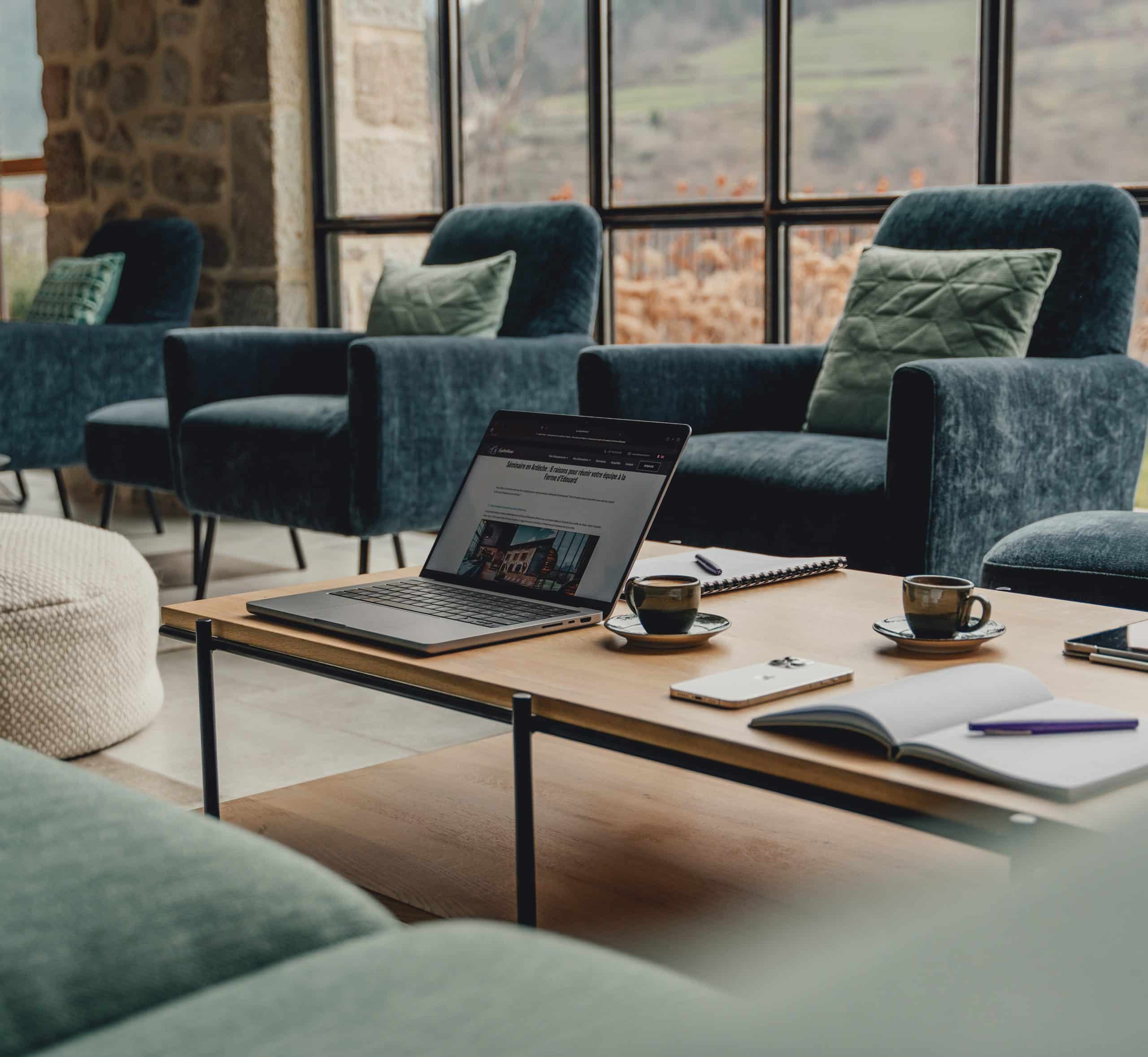 Ordinateur portable ouvert affichant "Séminaire en Ardèche : 8 raisons pour réunir votre équipe à la Ferme d'Édouard". Il est posé sur une table basse en bois, avec des tasses et carnets, dans un salon aux fauteuils bleus face à de grandes fenêtres.