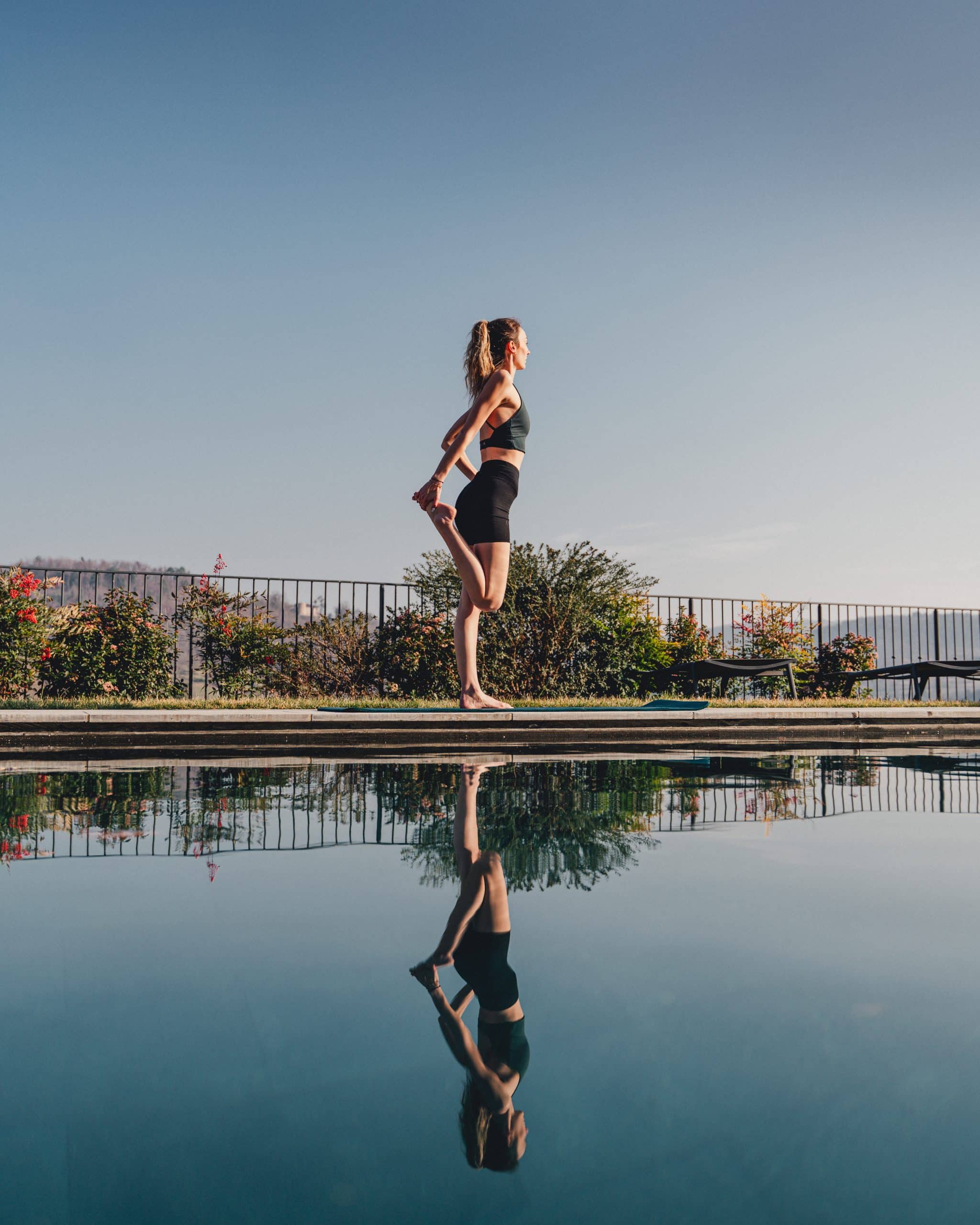 Une jeune femme étire son quadriceps droit, debout en bord de piscine. Un ciel clair et un paysage vallonné encadrent son reflet clair dans l'eau calme.
