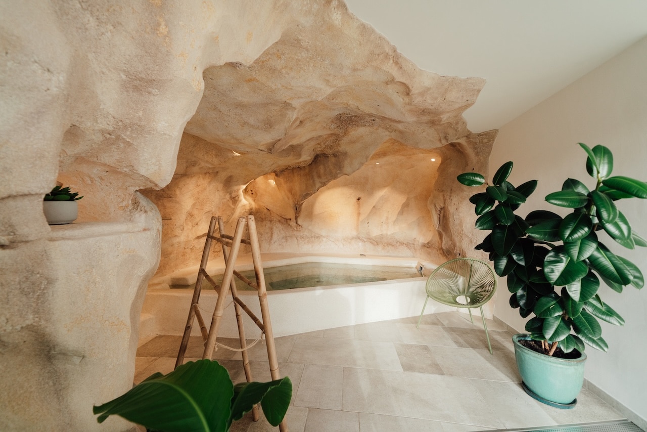 Jacuzzi creusé dans une grotte rocheuse artificielle, entouré de plantes vertes, d'une échelle en bambou et d'un fauteuil moderne, créant un espace spa aux tons clairs.
