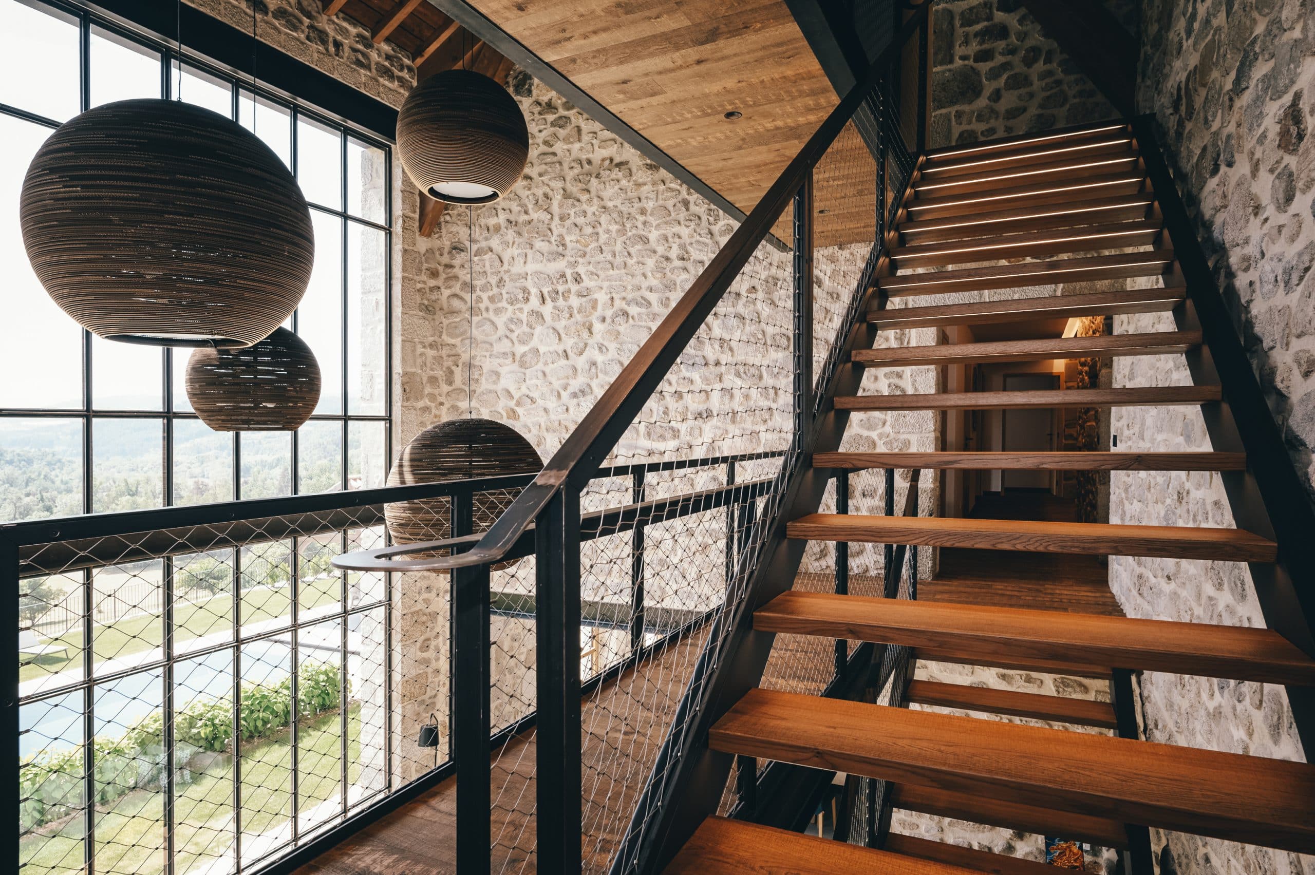 Un escalier moderne en bois clair, éclairé, monte le long d'un mur en pierres et de grandes baies vitrées offrant une vue sur un paysage verdoyant, sous des luminaires sphériques en carton.