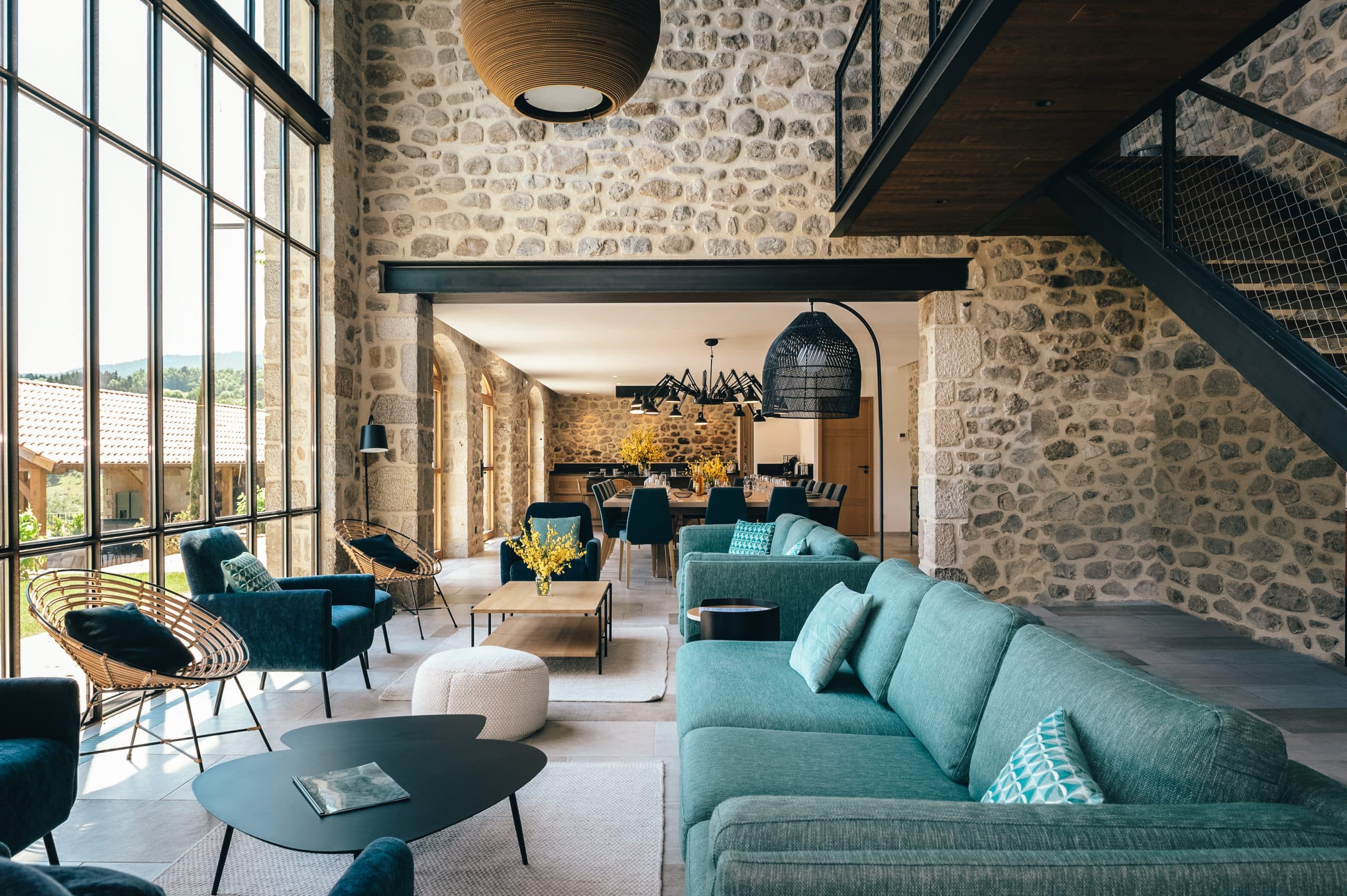 Intérieur moderne, espace ouvert salon-salle à manger avec mobilier turquoise et rotin. Murs en pierre, grandes baies vitrées sur la nature. Mezzanine avec escalier métallique.