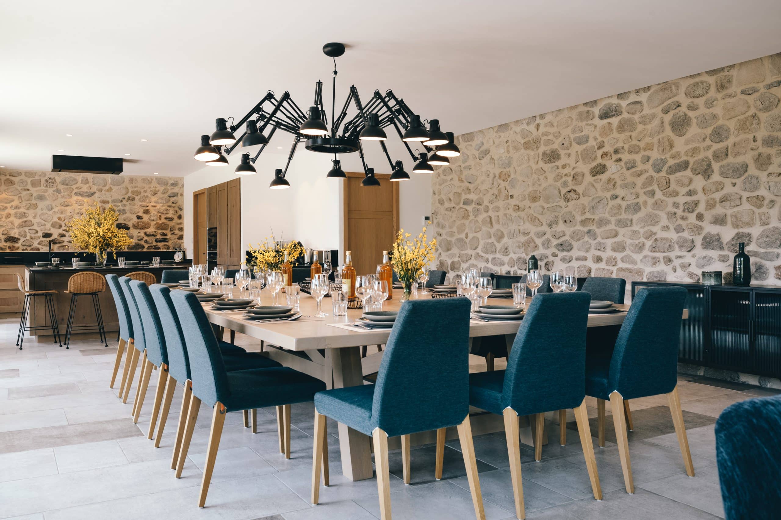 Une longue table de salle à manger dressée, entourée de chaises bleues, est surmontée d'un lustre araignée noir dans une pièce aux murs de pierre et sol carrelé.