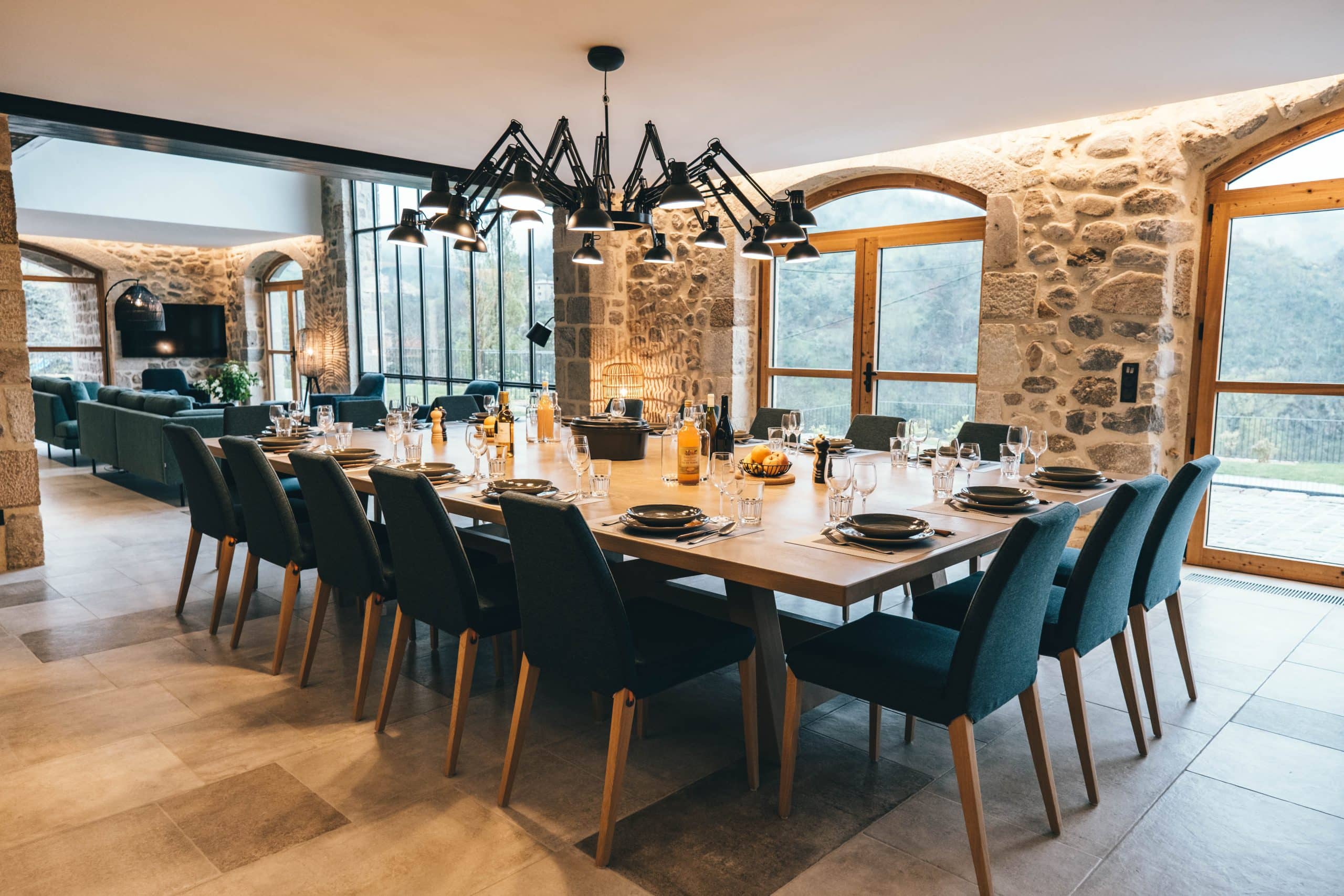 Longue table en bois dressée pour dix-huit personnes avec vaisselle et boissons. Elle domine une grande pièce aux murs en pierres, larges fenêtres et un coin salon attenant.