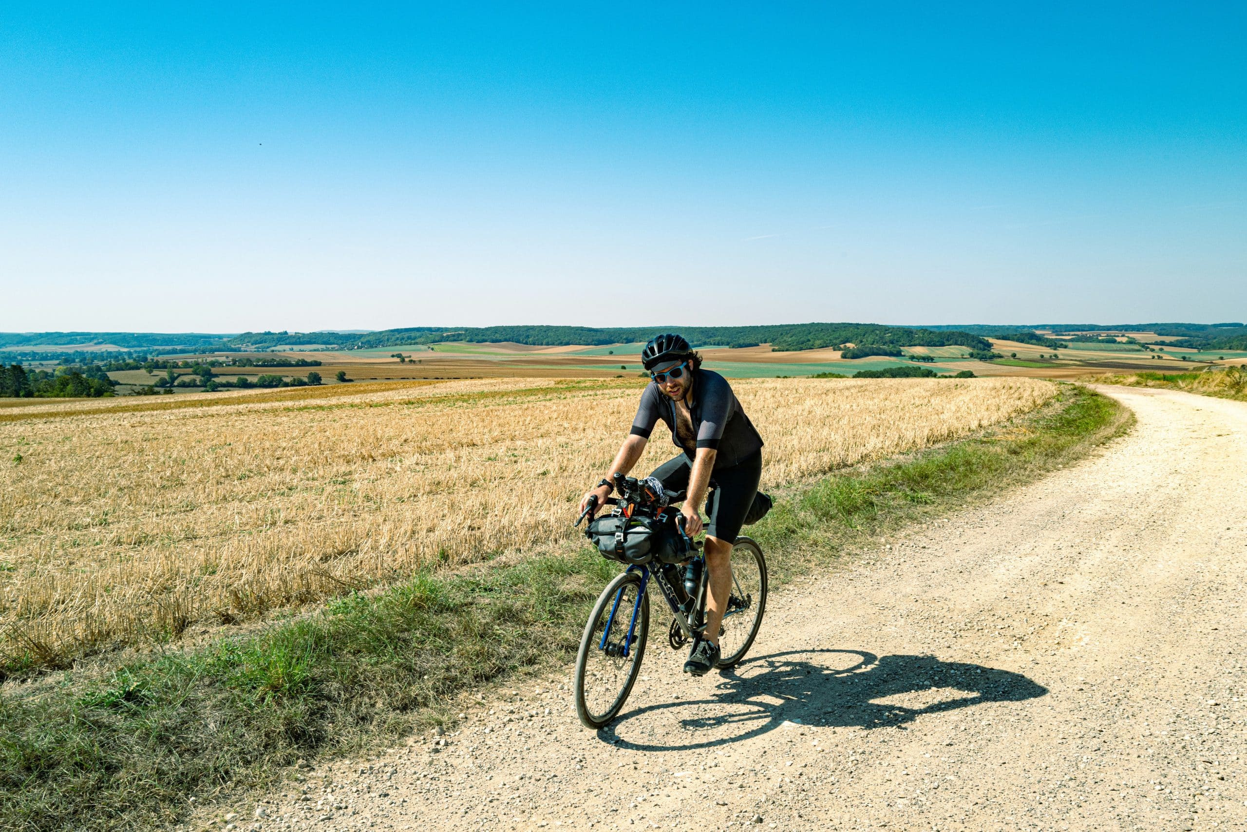 Un homme équipé d'un casque et de lunettes fait du vélo de randonnée sur un chemin de terre sinueux, entre des champs moissonnés et des collines verdoyantes sous un ciel bleu.