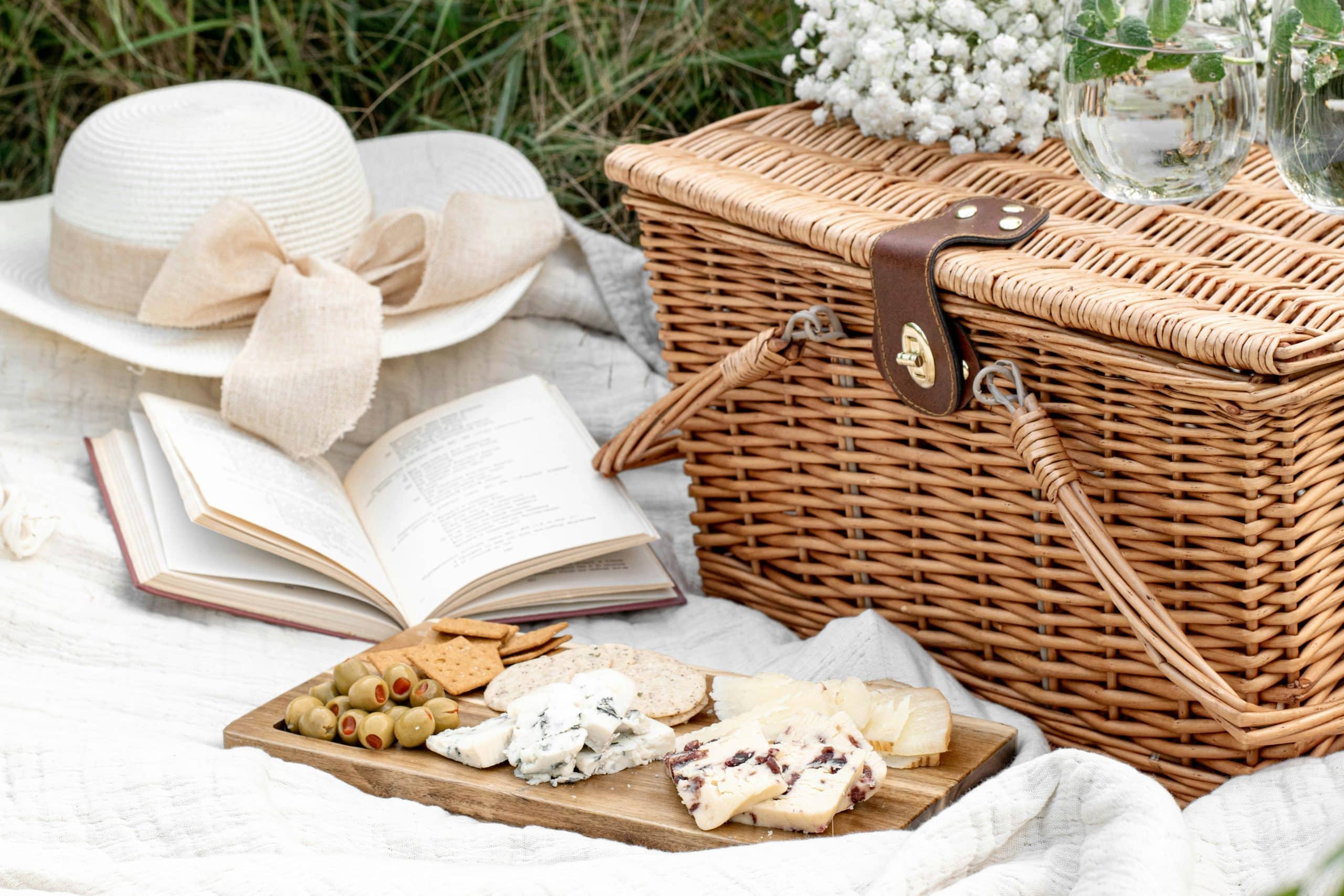 Panier de pique-nique en osier, chapeau, livre ouvert et planche de fromages, olives, crackers sont disposés sur une couverture blanche dans l'herbe. Des verres mentholés et des fleurs complètent la scène.