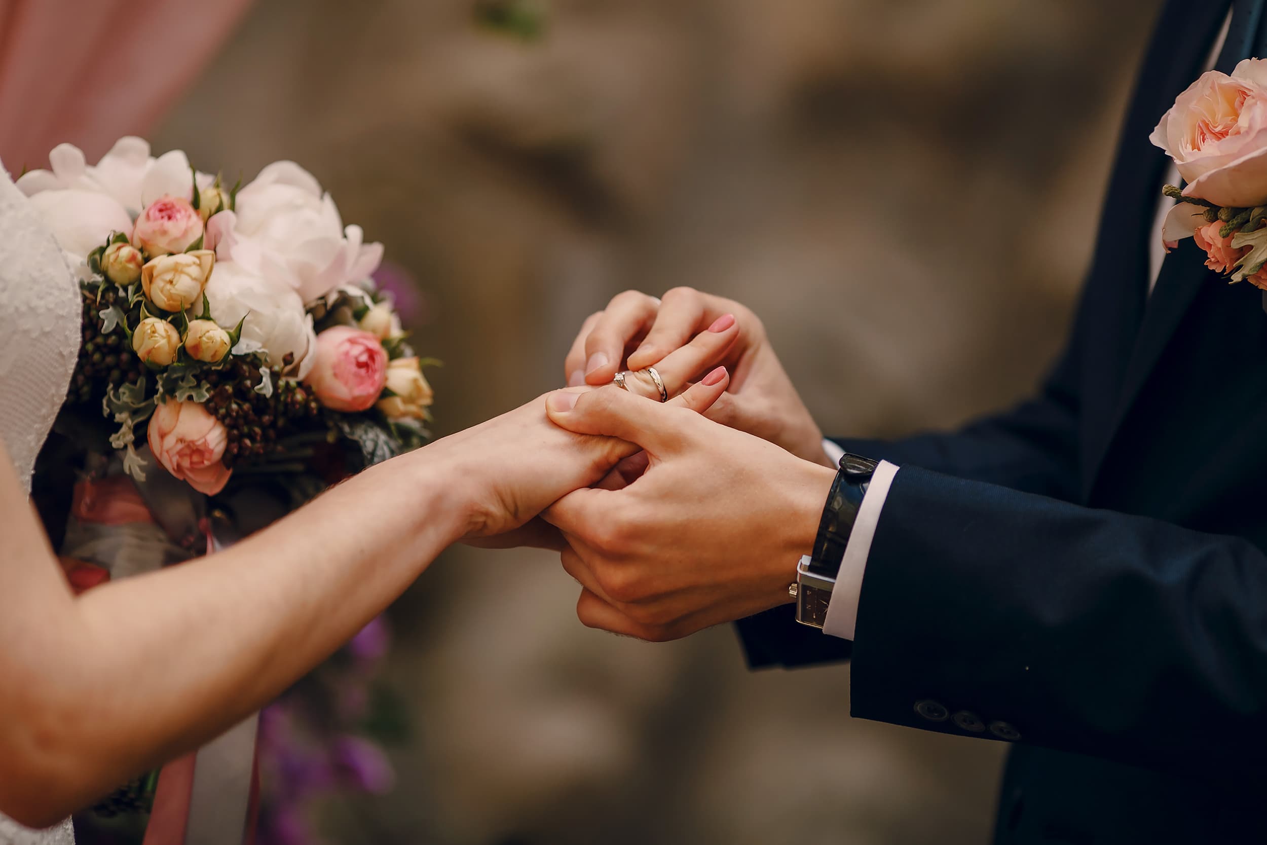 Une main masculine passe une alliance au doigt d'une main féminine manucurée, avec un bouquet de mariage floral pastel en arrière-plan flou.