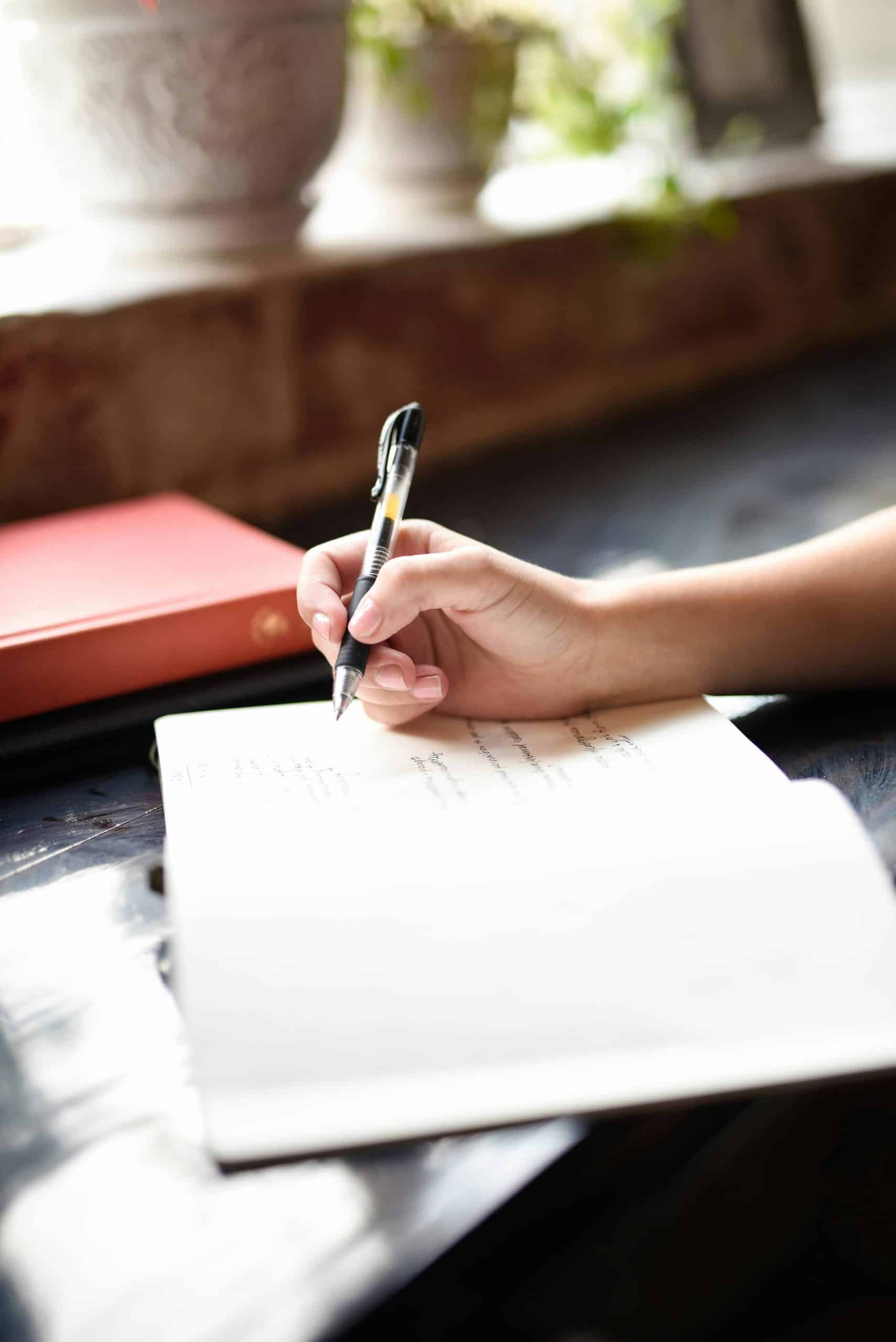 Une main écrit avec un stylo sur un cahier ouvert, posé sur une surface sombre. Un livre rouge est visible à gauche, avec un arrière-plan flou et lumineux.