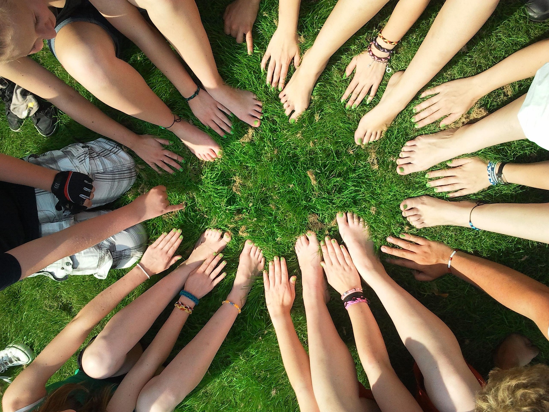 De nombreuses personnes sont allongées, formant un cercle avec leurs bras et jambes convergeant vers un centre sur l'herbe verte. Leurs mains et pieds, aux ongles vernis vert clair, se touchent.