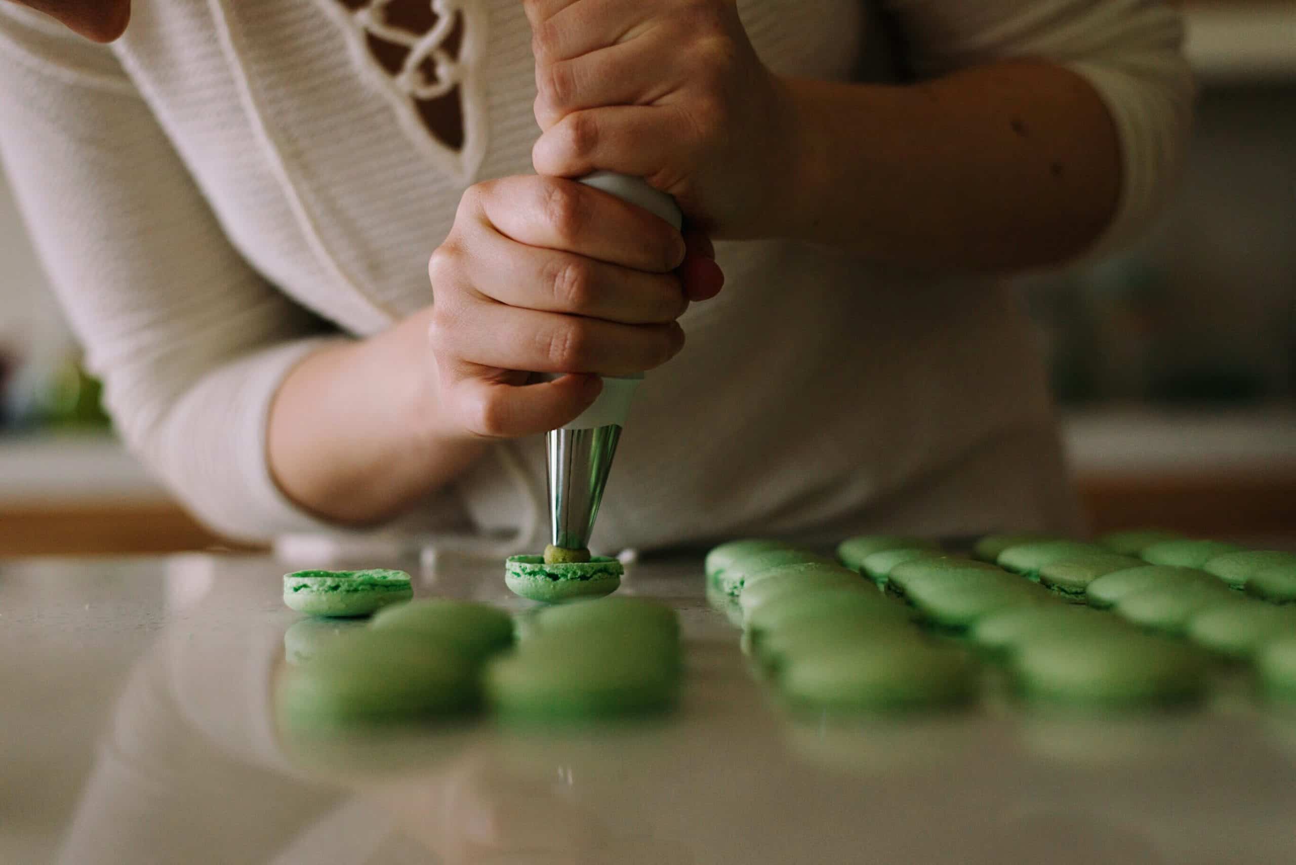 Des mains remplissent des coques de macarons verts avec une poche à douille, sur une surface claire. De nombreux macarons verts alignés sont visibles à droite.