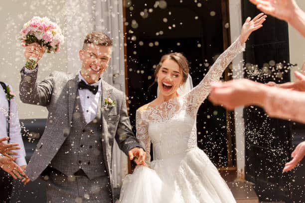 Les jeunes mariés, souriants et joyeux, sortent d'une porte, sous une pluie de riz/confettis lancés par des invités, le marié tenant un bouquet, la mariée les bras levés.