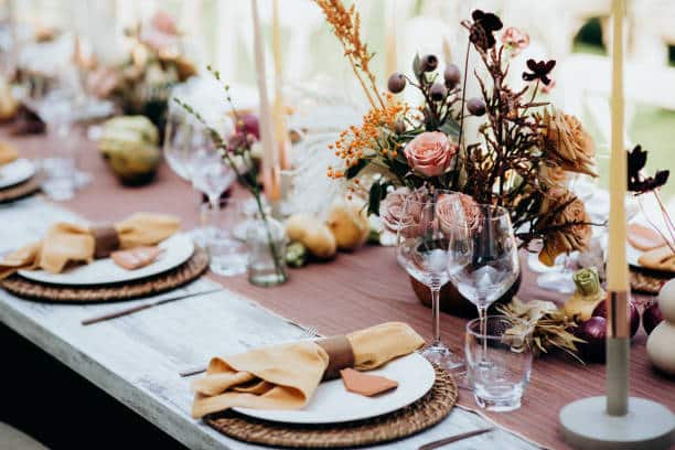 Une longue table en bois clair est dressée avec des sets, assiettes, serviettes ocres et verres. Des centres de table fleuris de roses et branchages, bougies et fruits la décorent élégamment.