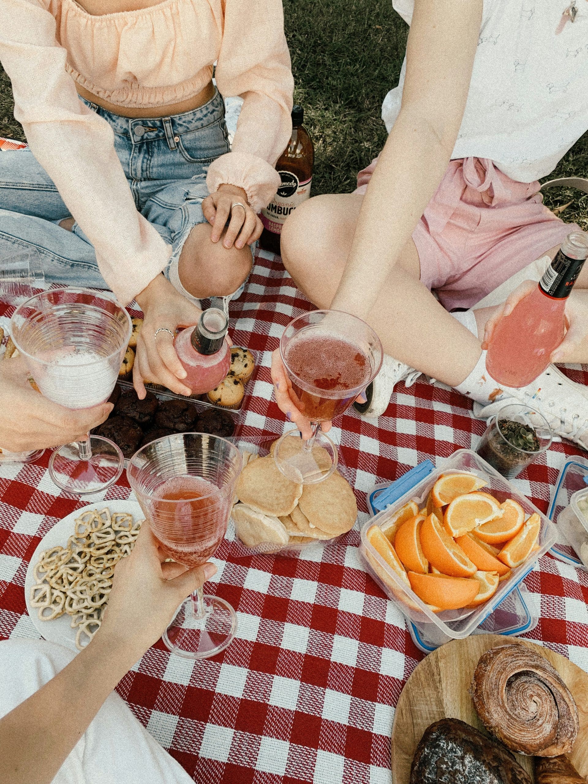 Plusieurs personnes partagent un pique-nique, tenant des boissons et des snacks variés sur une nappe à carreaux rouges et blancs, posée sur l'herbe. Texte sur une bouteille : "medy organic OMBUC RASPBE LEMON".