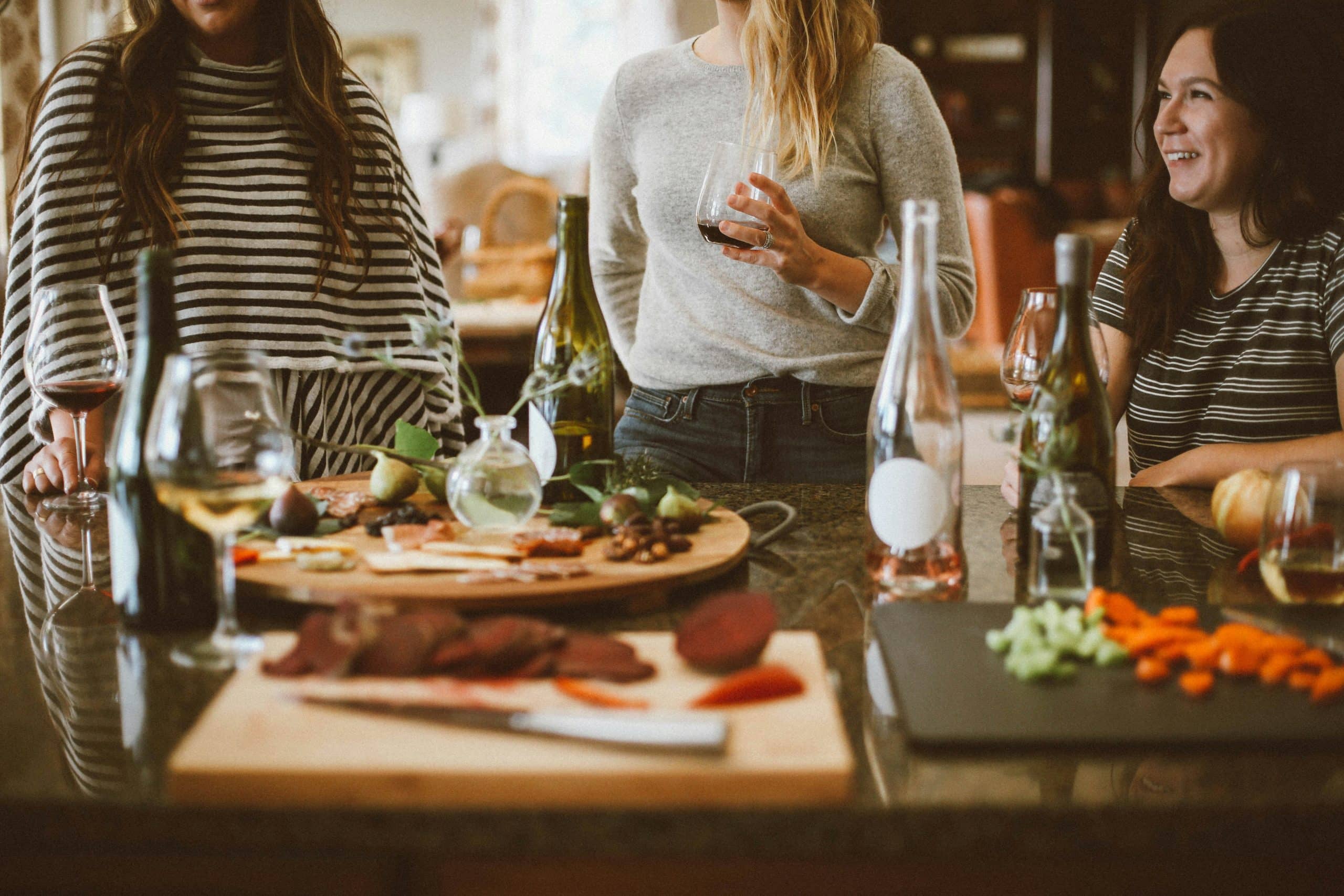 Trois femmes se réunissent autour d'un comptoir. Elles boivent du vin parmi des plateaux de fromages, charcuteries, légumes et fruits secs, dans une ambiance conviviale.
