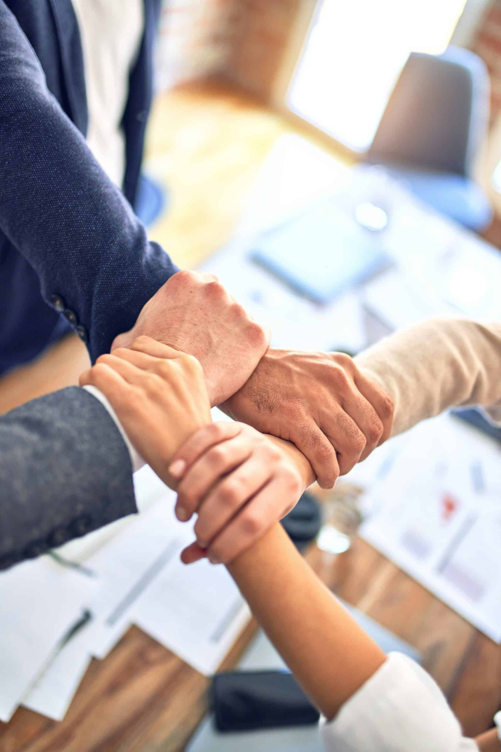 Quatre avant-bras et mains de personnes diverses s'enlacent en cercle sur une table de bureau avec des documents flous, évoquant la collaboration.