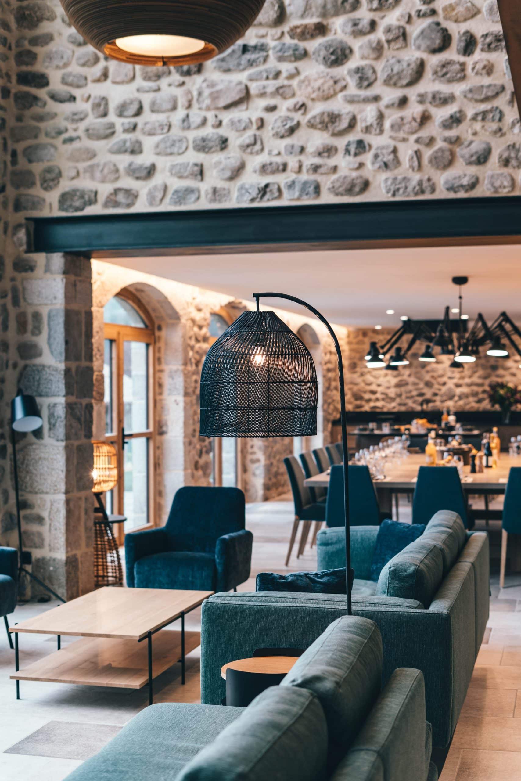 Salon moderne avec canapés et fauteuils bleu-vert, lampadaire design. Murs de pierre, portes cintrées ouvrant sur une salle à manger aux tables dressées.