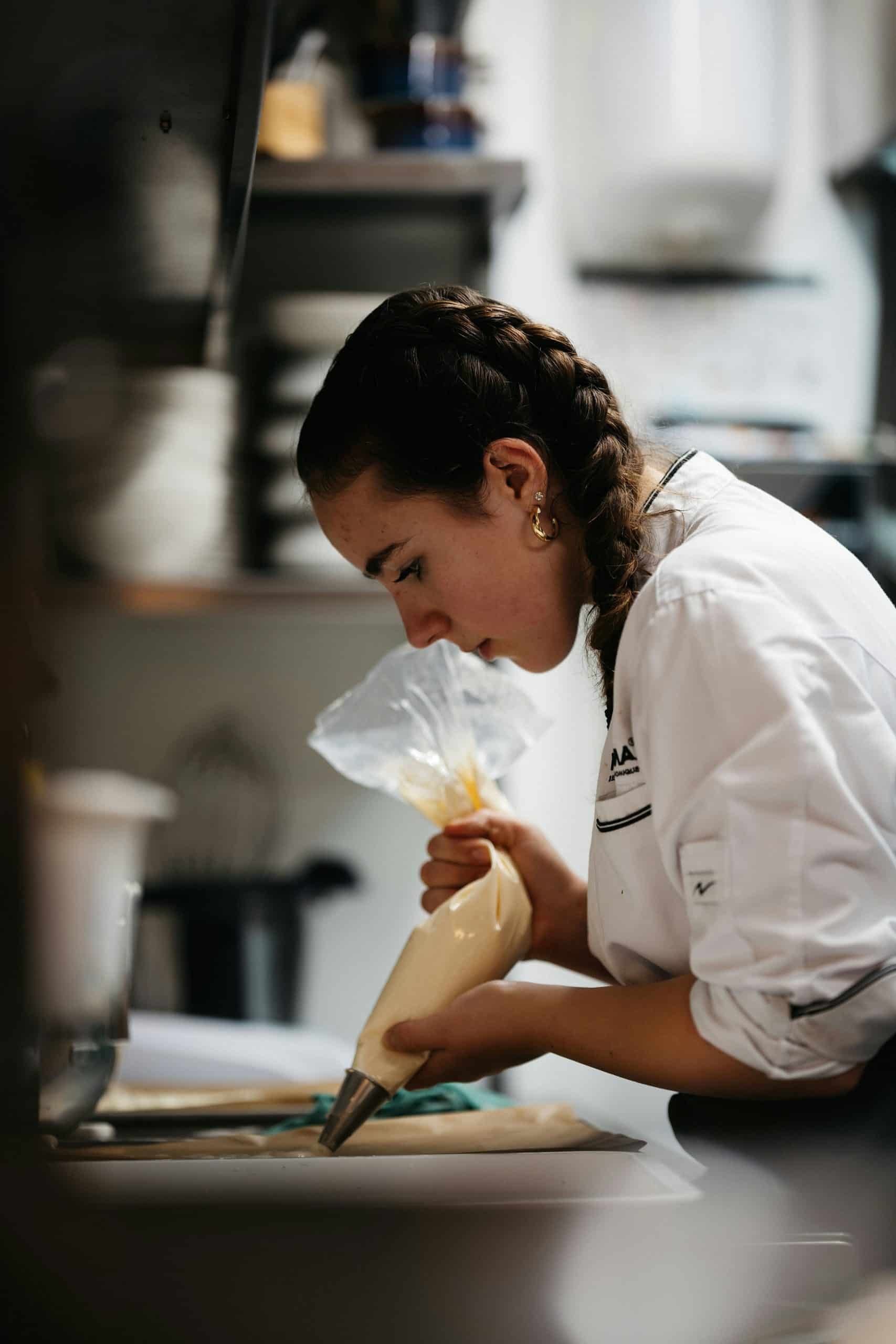 Une jeune cheffe, tresses brunes et veste blanche marquée "AA MONIQUE", poche une crème jaune pâle avec concentration sur un plan de travail en cuisine professionnelle.