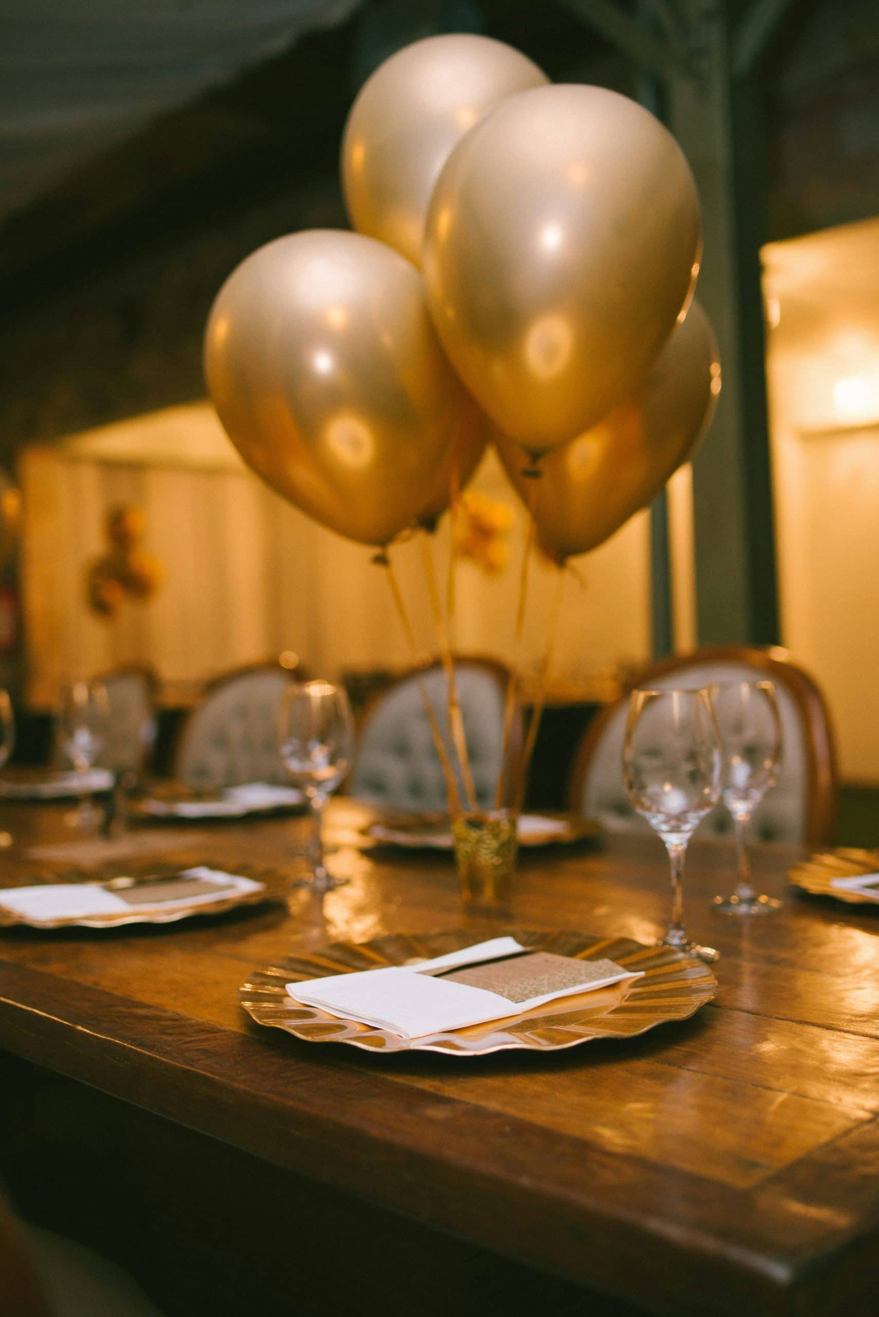 Des ballons dorés flottent au-dessus d'une longue table en bois dressée avec des assiettes dorées, des serviettes et des verres, dans une ambiance chaleureuse de salle à manger.