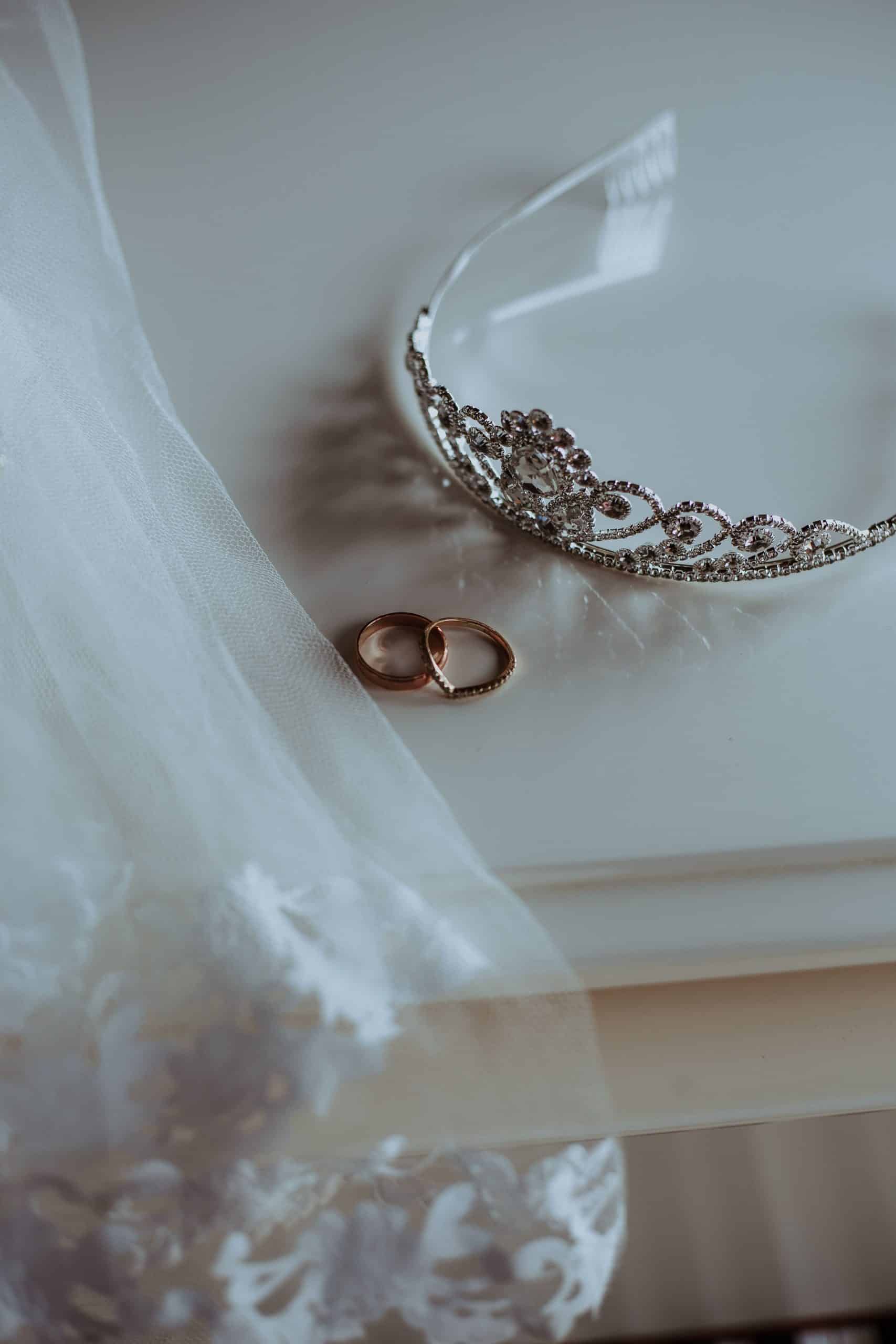 Une tiare ornée de pierres et deux alliances dorées sont posées sur une surface claire et réfléchissante. Un voile de mariée blanc, finement brodé, est drapé à côté d'elles.