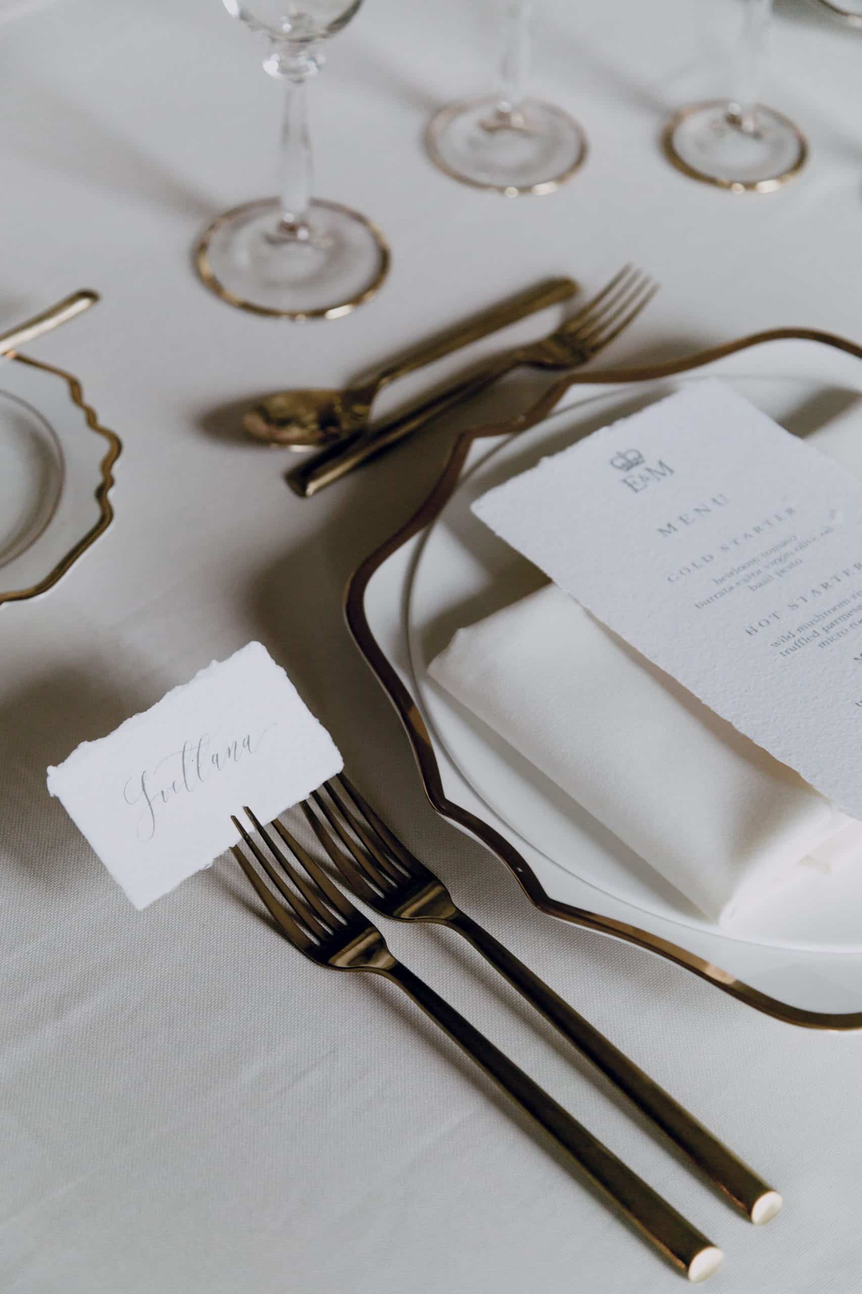 Un couvert de table élégant est dressé sur une nappe claire, avec des assiettes bordées d'or, des couverts dorés et une serviette roulée. Un menu affiche "E & M", "MENU", "COLD STARTER" (heirloom tomato, burrata extra virgin olive oil, basil pesto), "HOT STARTER" (wild mushroom ra, truffled parmesan, micro roquett). Une carte de nom "Svetlana" est posée sur une fourchette.