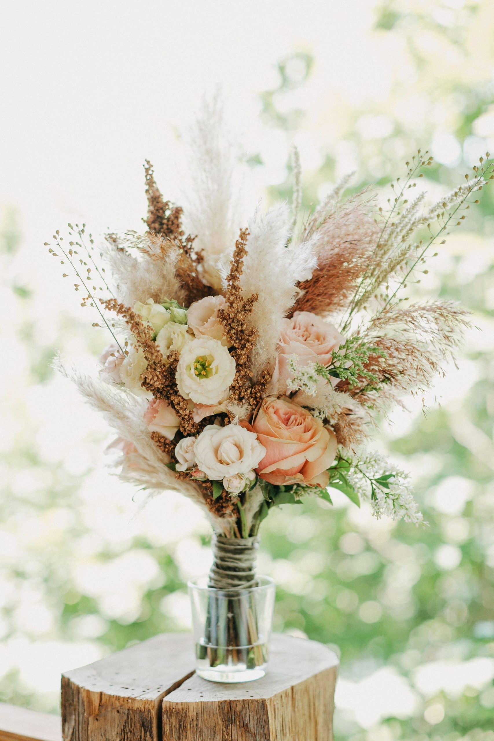 Un bouquet de roses pêche, lisianthus crème et herbes de la pampa est posé dans un verre sur une bûche en bois, devant un arrière-plan vert flou.