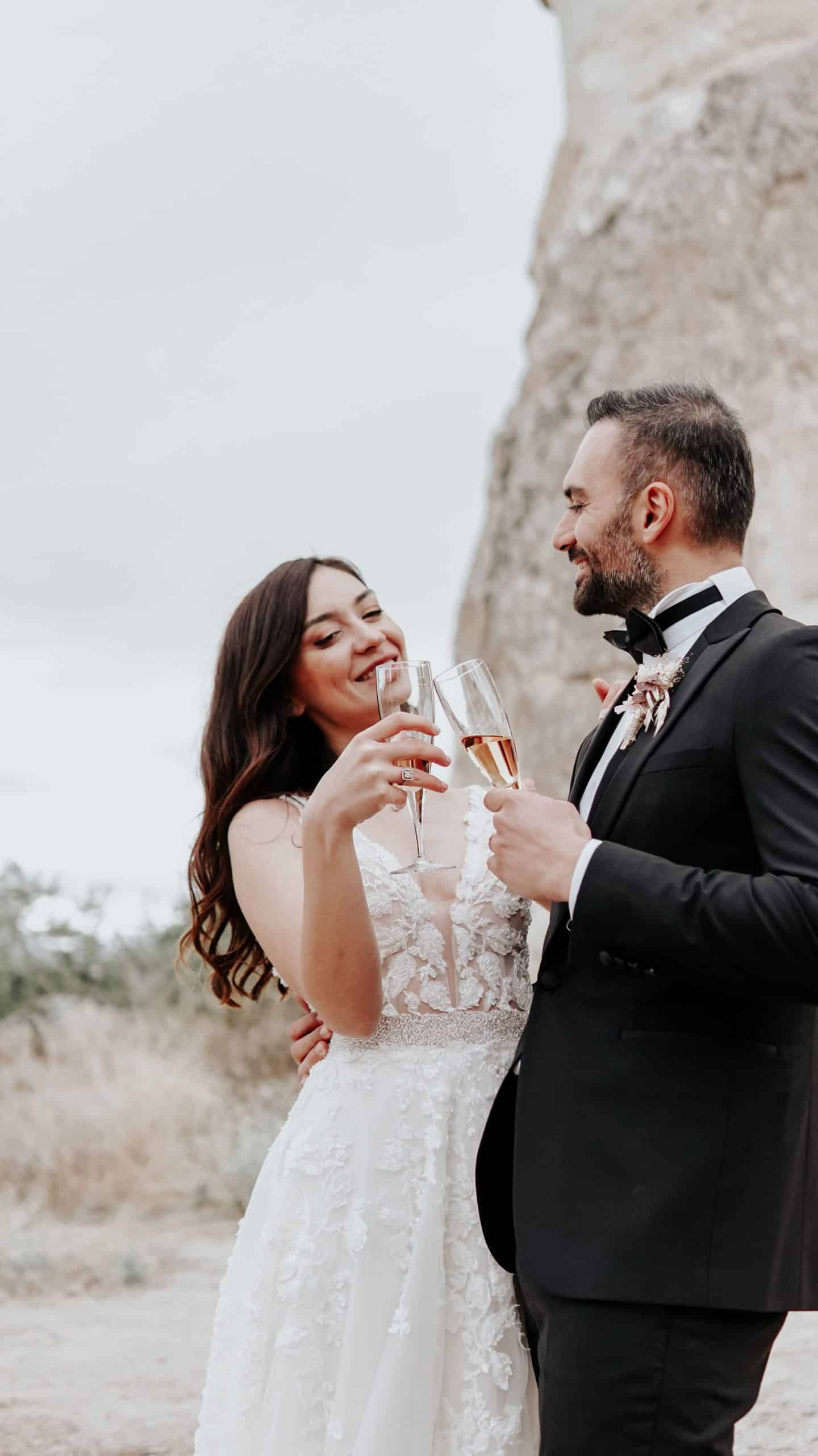 Un couple, la mariée en robe blanche et le marié en smoking, lèvent des flûtes de champagne rosé en se souriant. Ils sont en extérieur devant une formation rocheuse et une végétation sèche.
