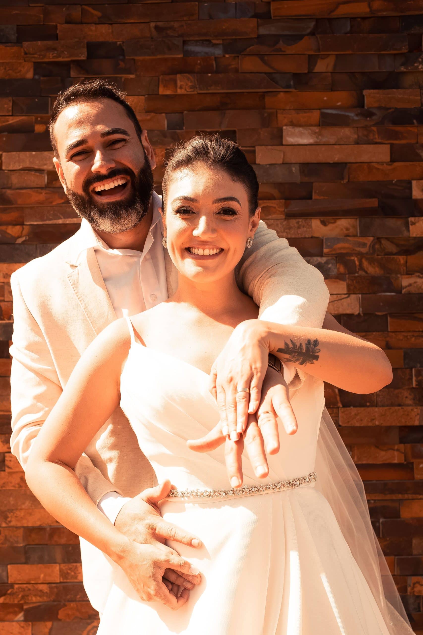 Un couple souriant, l'homme enlaçant la femme par-derrière, leurs mains superposées affichant des alliances, devant un mur de briques de pierre marron.