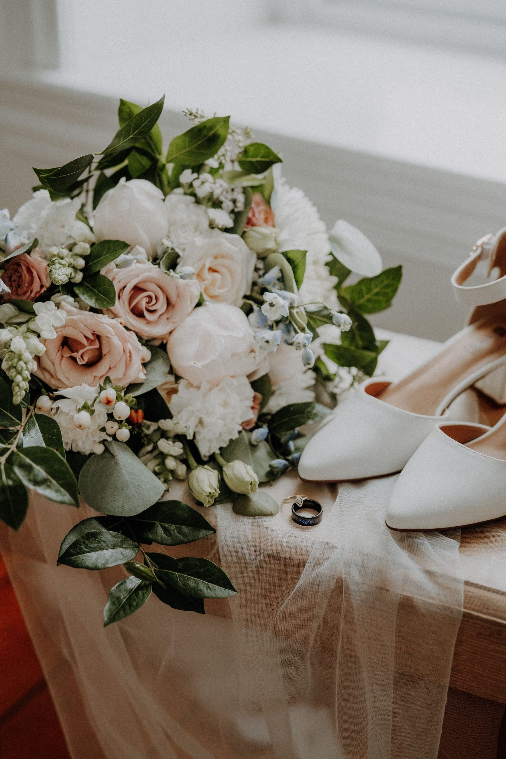 Un bouquet de mariage, des chaussures de mariée blanches et deux alliances sont posés sur un tissu clair drapé sur une surface en bois près d'une fenêtre.